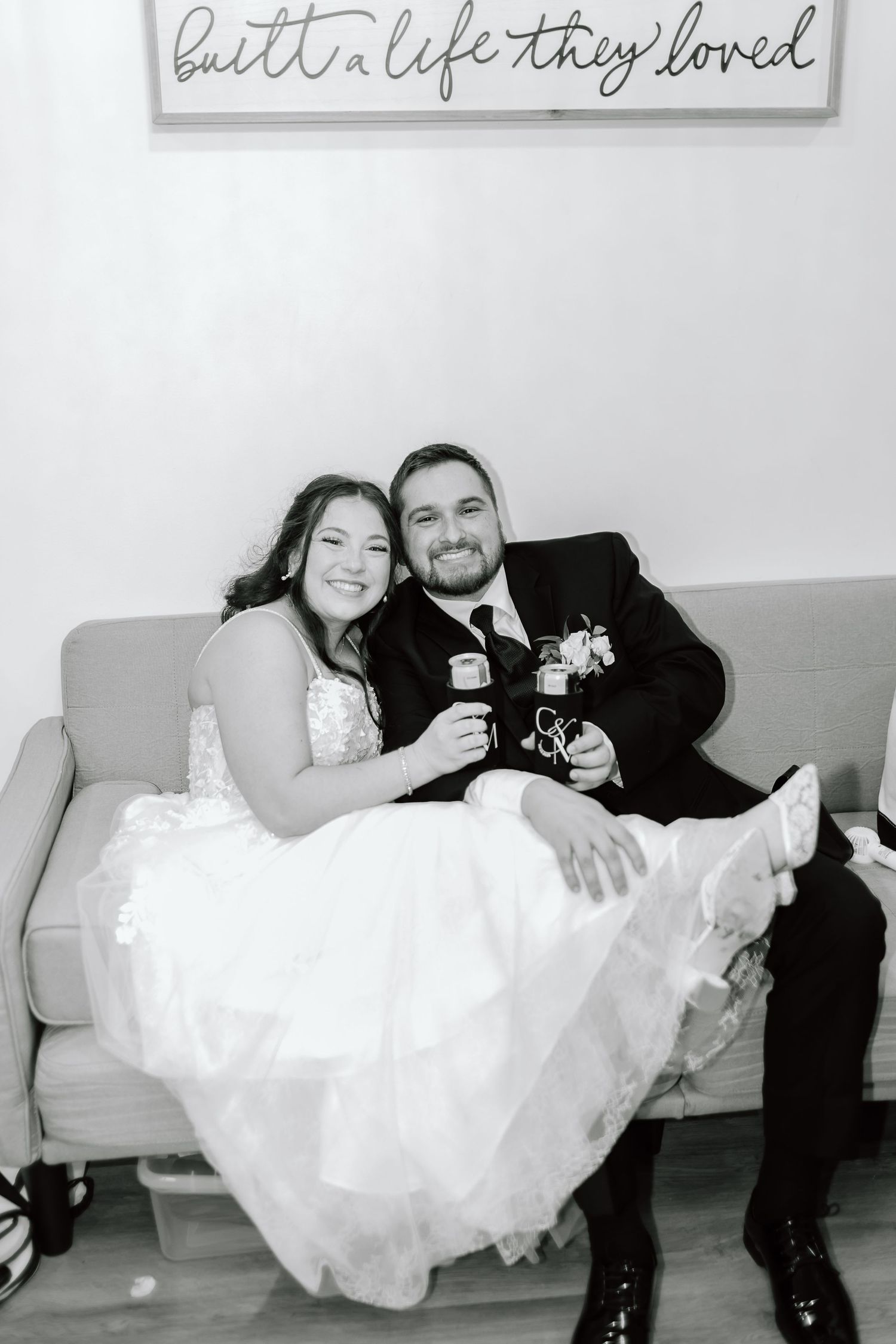 A bride and groom sit together on a gray couch, holding wine glasses beneath a decorative wall sign.