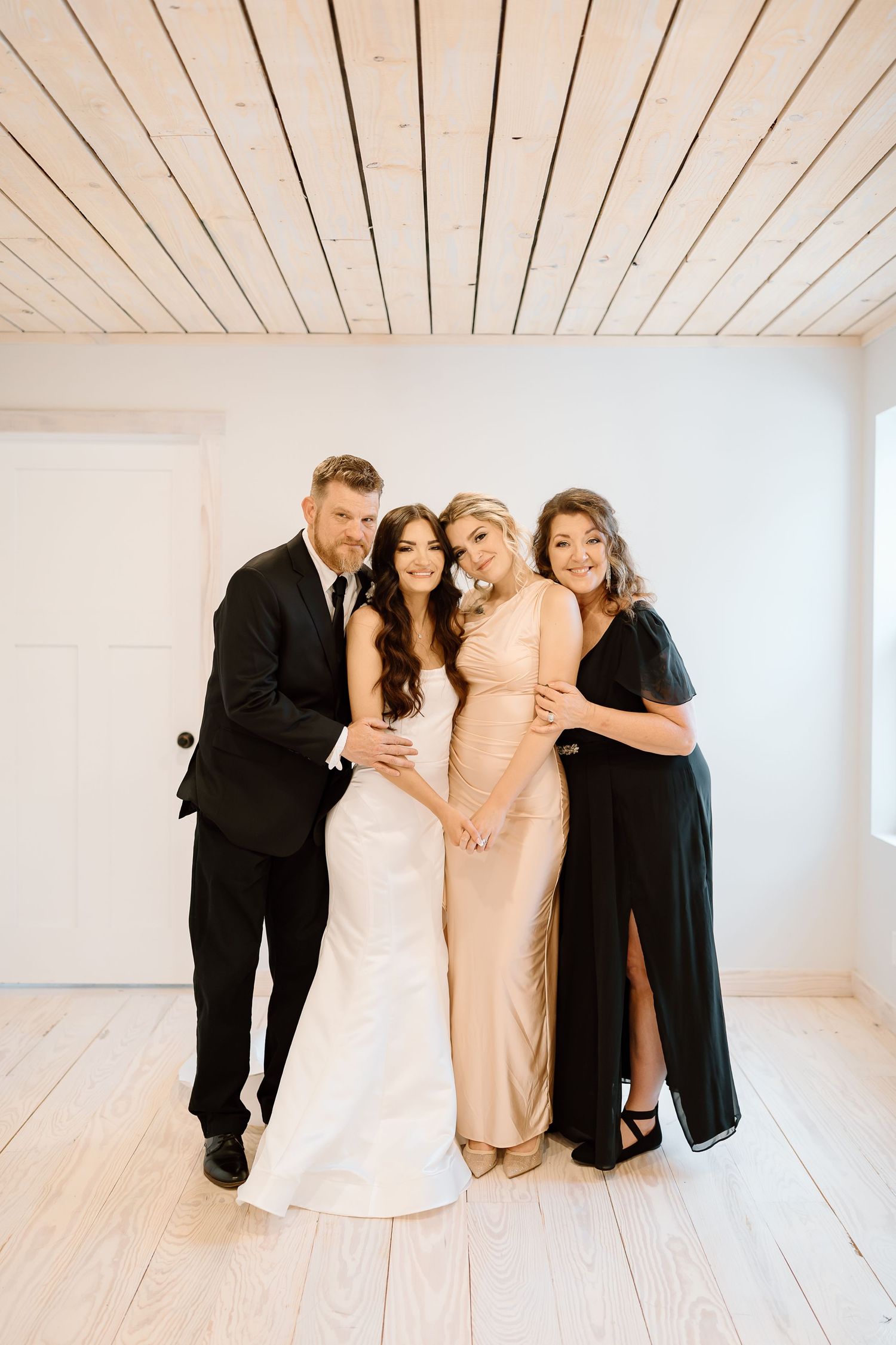 A group in formal attire shares a joyful moment together in a bright white studio space with wooden floors.