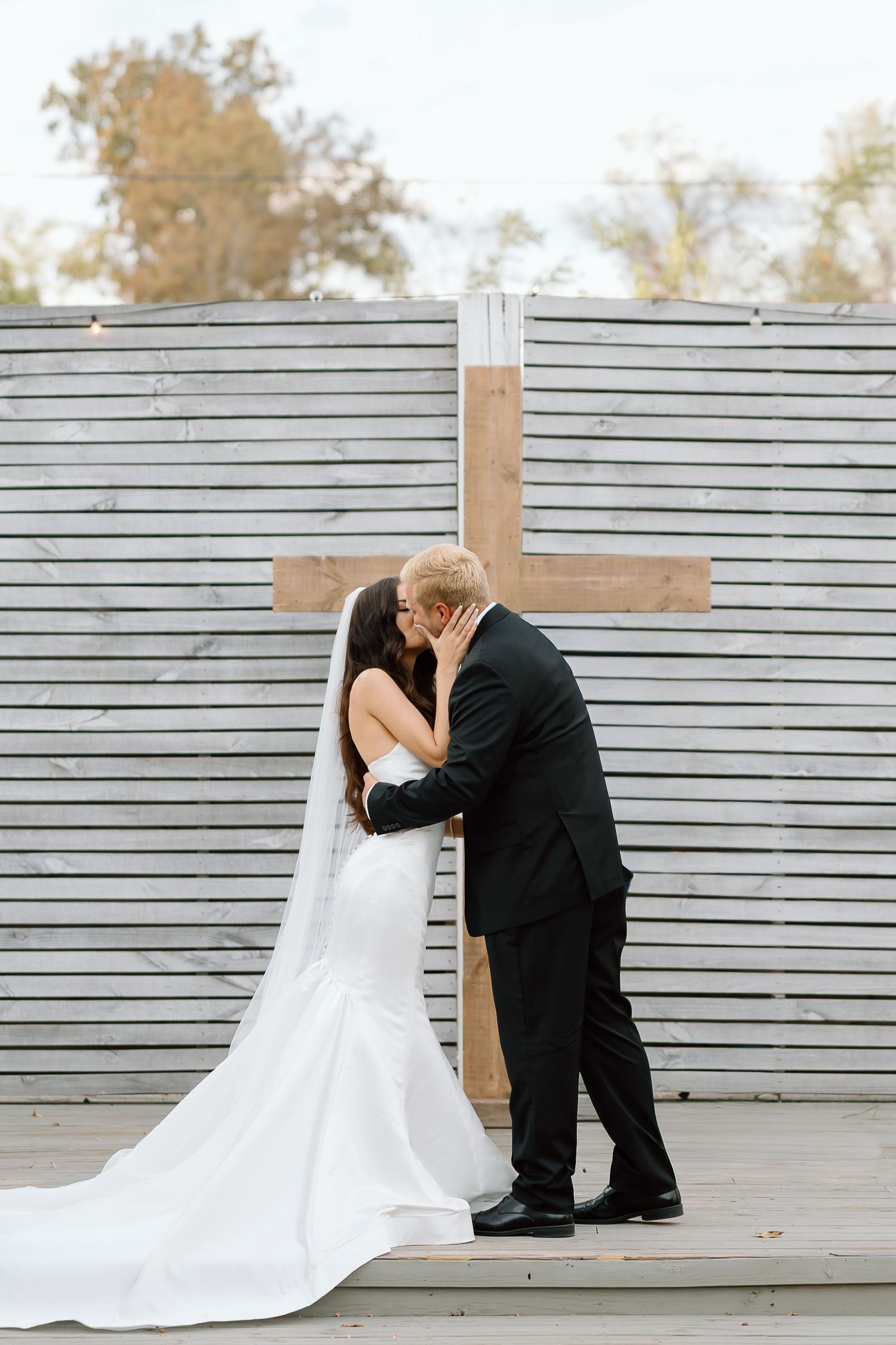 A romantic wedding moment captured in front of a wooden cross and grey slatted wall backdrop.