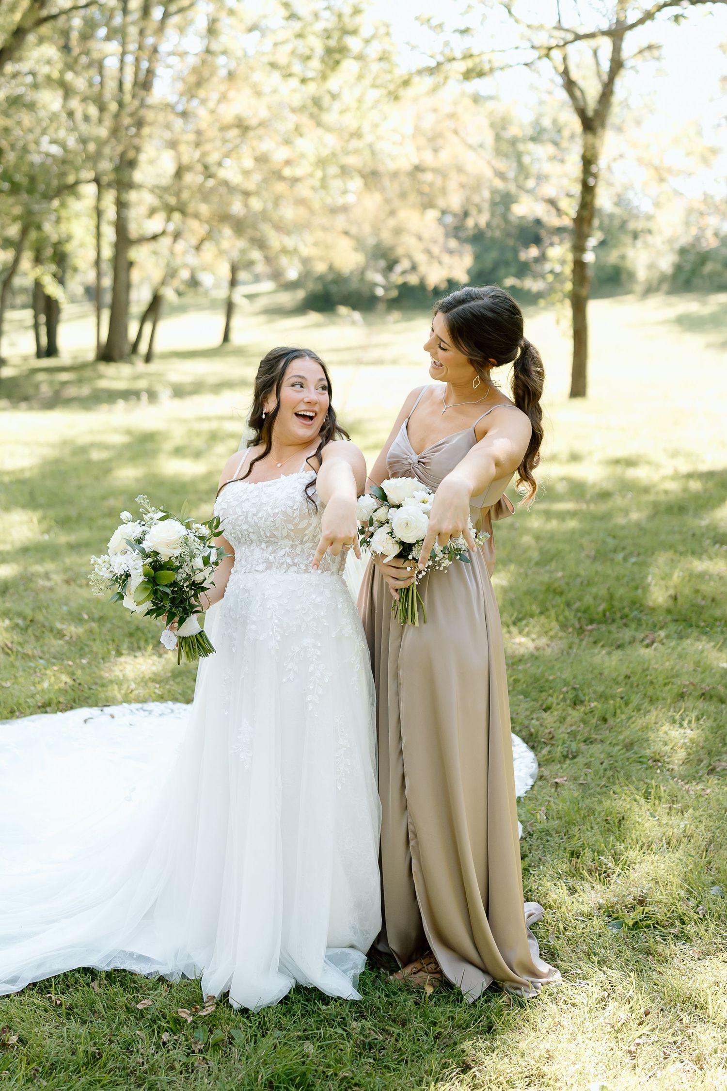 Two wedding attendees sit together on grass in a sunlit park, sharing joyful laughter while holding floral bouquets.