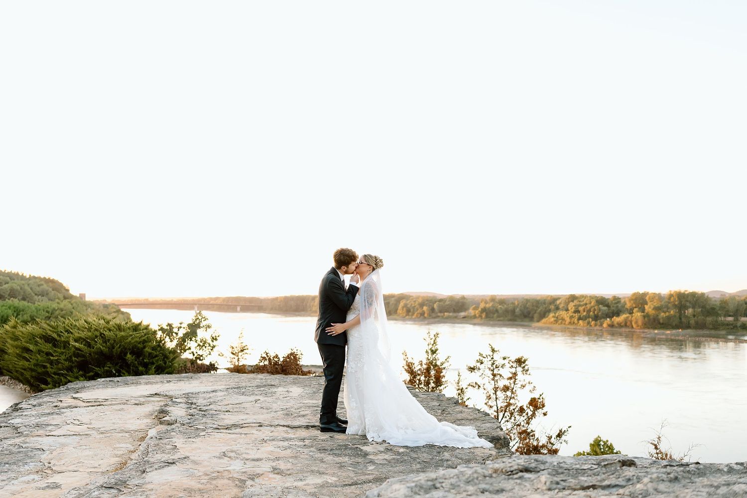 A couple stands together in formal wedding attire on a rocky shore with a scenic lake and autumn trees in the background.