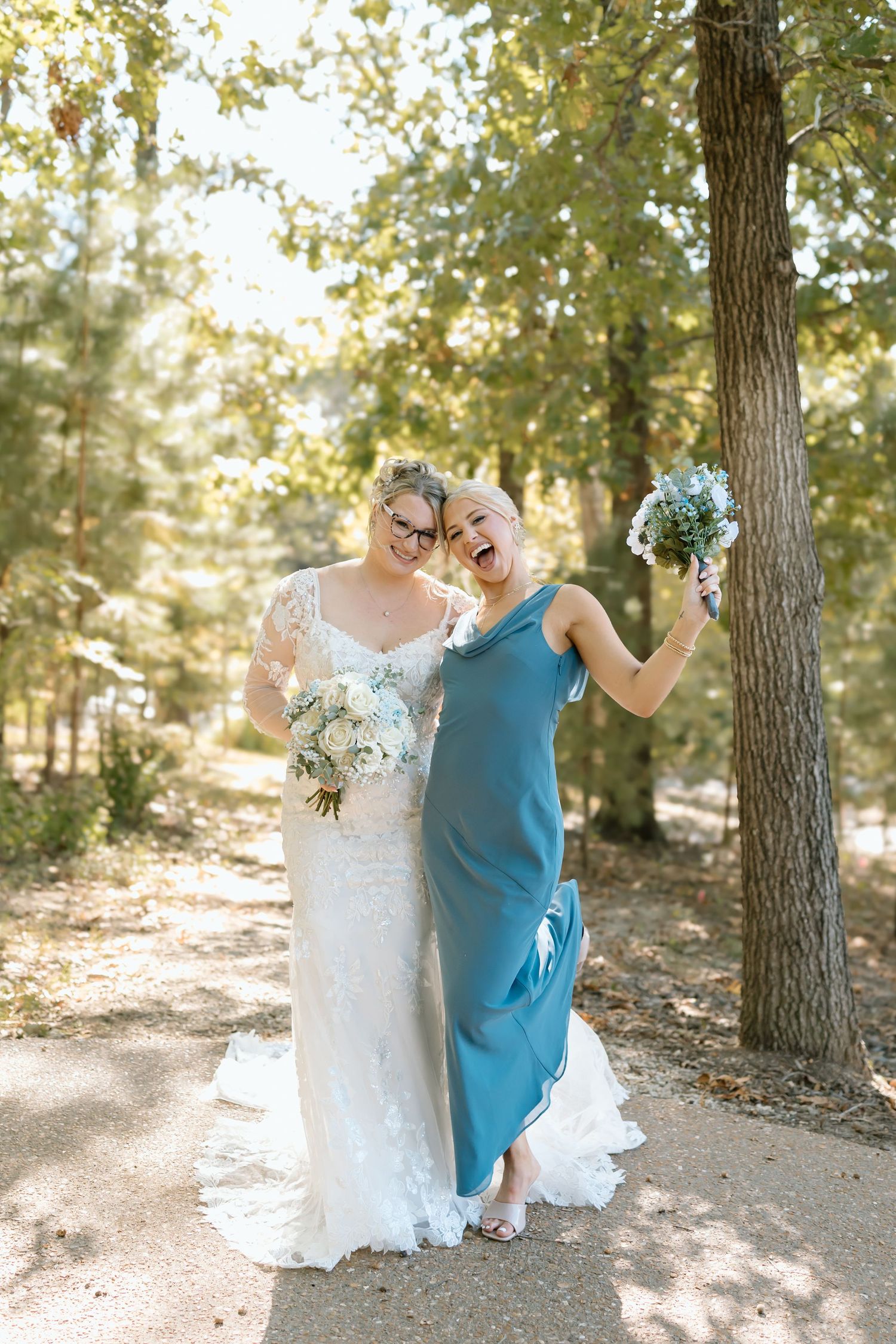 Two people in wedding attire dance and celebrate together on a wooded path surrounded by tall trees.