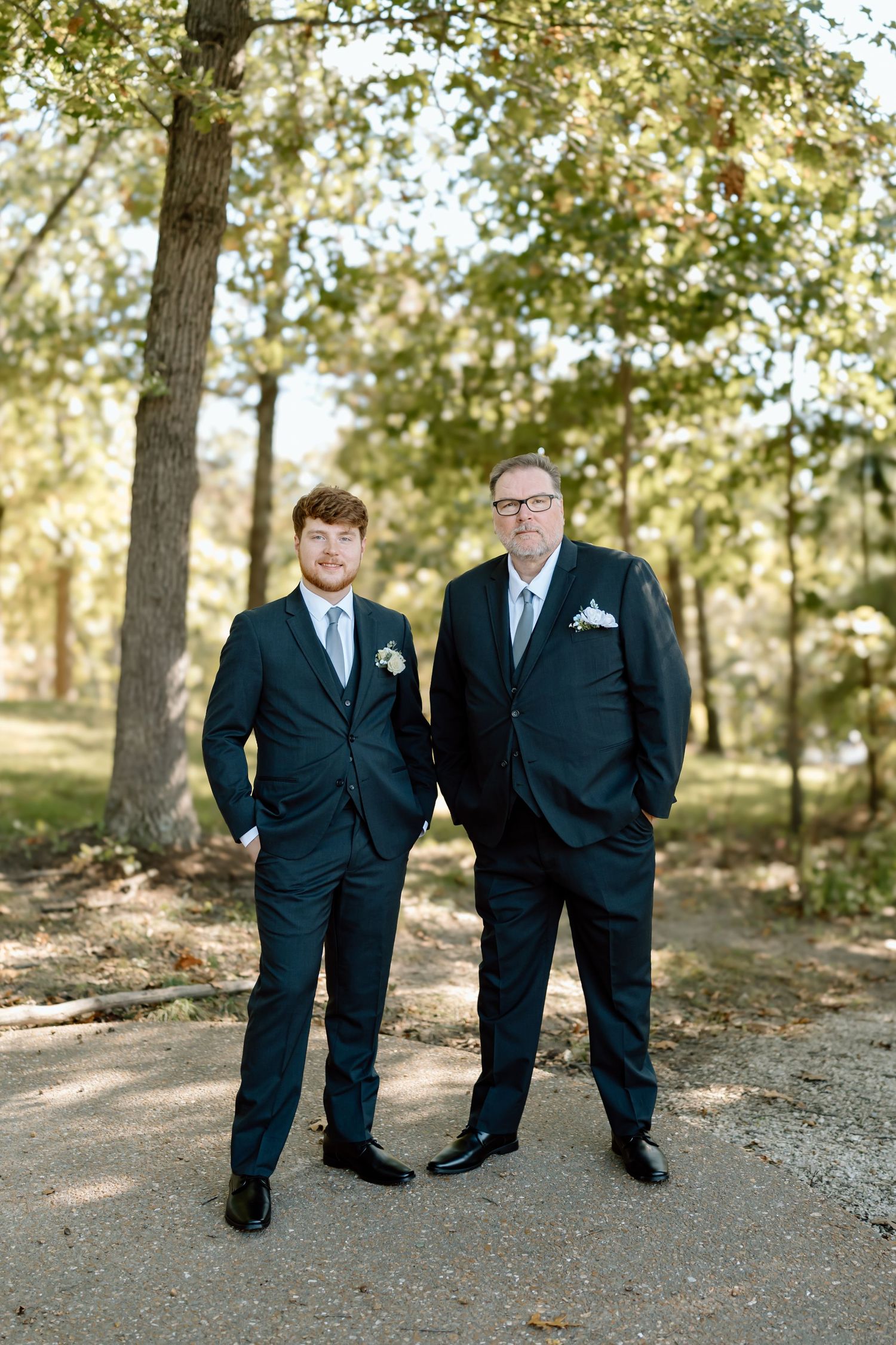 Two people in dark suits standing together on a wooded path in autumn.