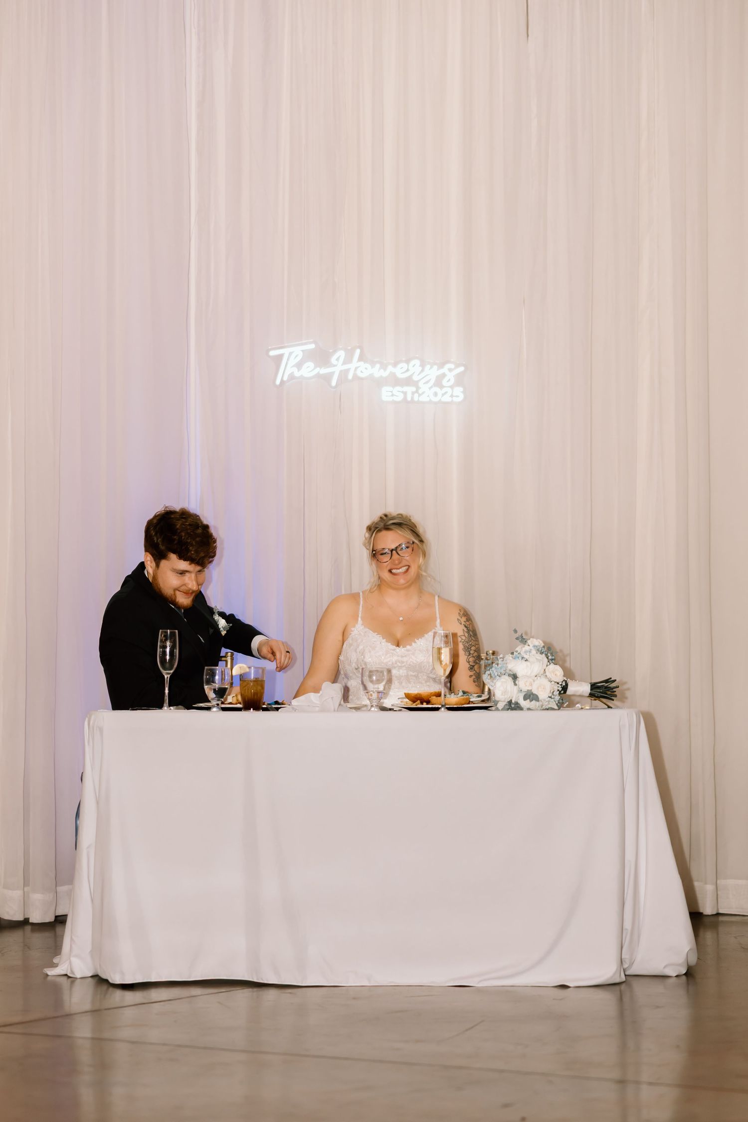Couple and wedding party members seated at elegant white head table during reception.