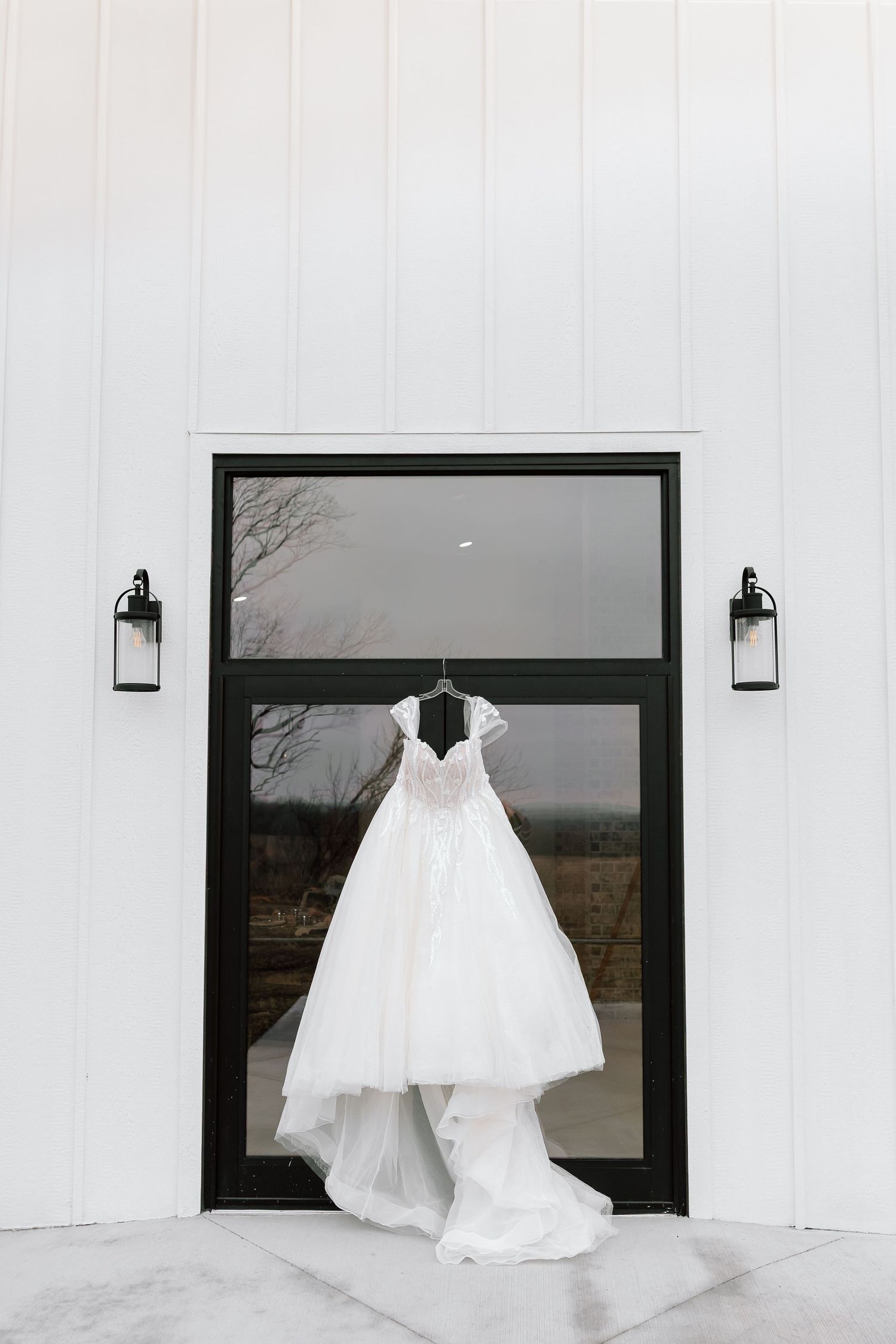 A white wedding dress hangs elegantly against a modern black-framed window on a white exterior wall.