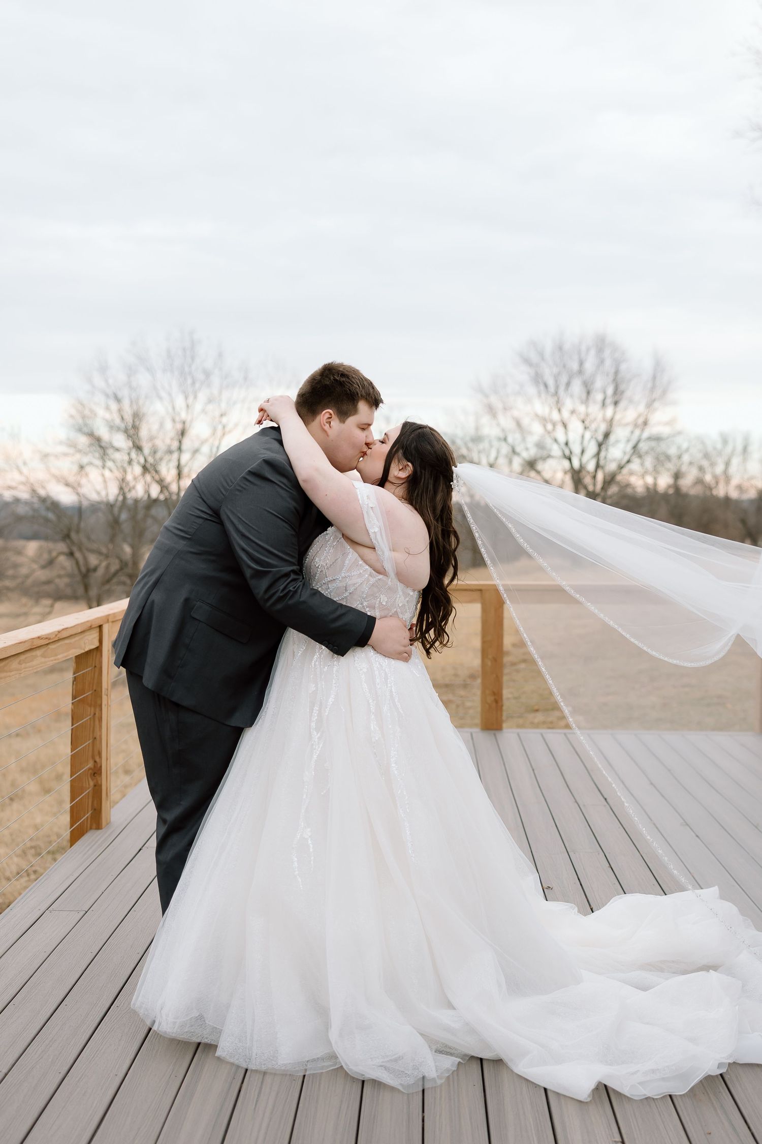 A romantic wedding couple shares an intimate dip kiss on a wooden deck surrounded by winter trees.