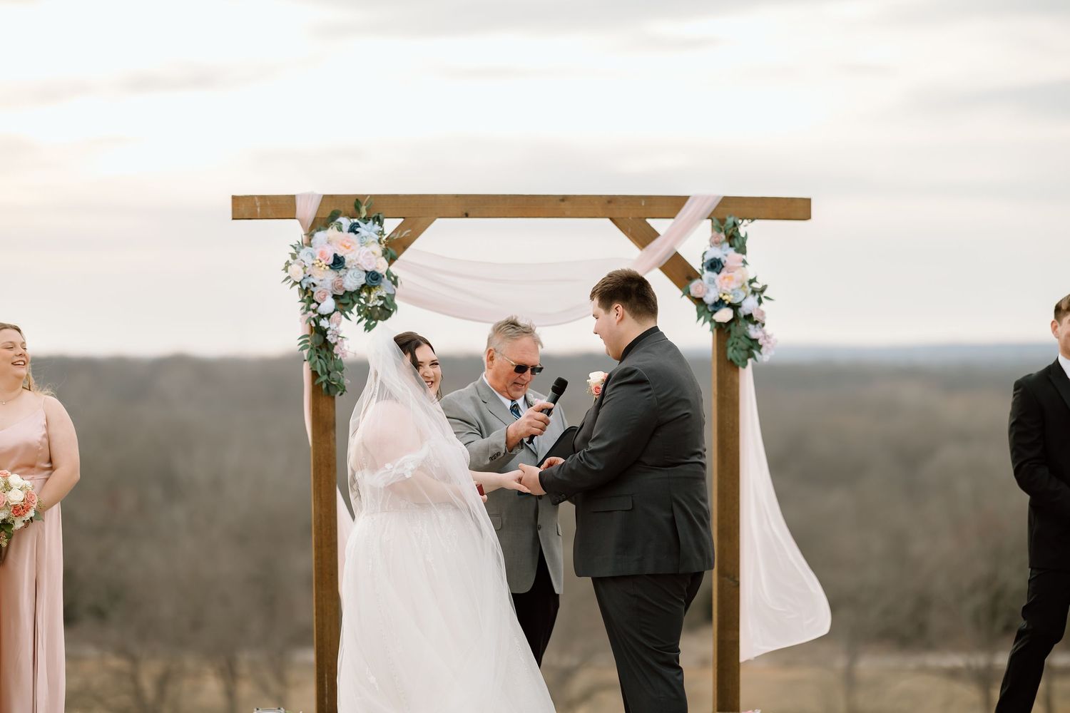 A couple exchanges vows during an outdoor wedding ceremony beneath a wooden cross with mountain views.