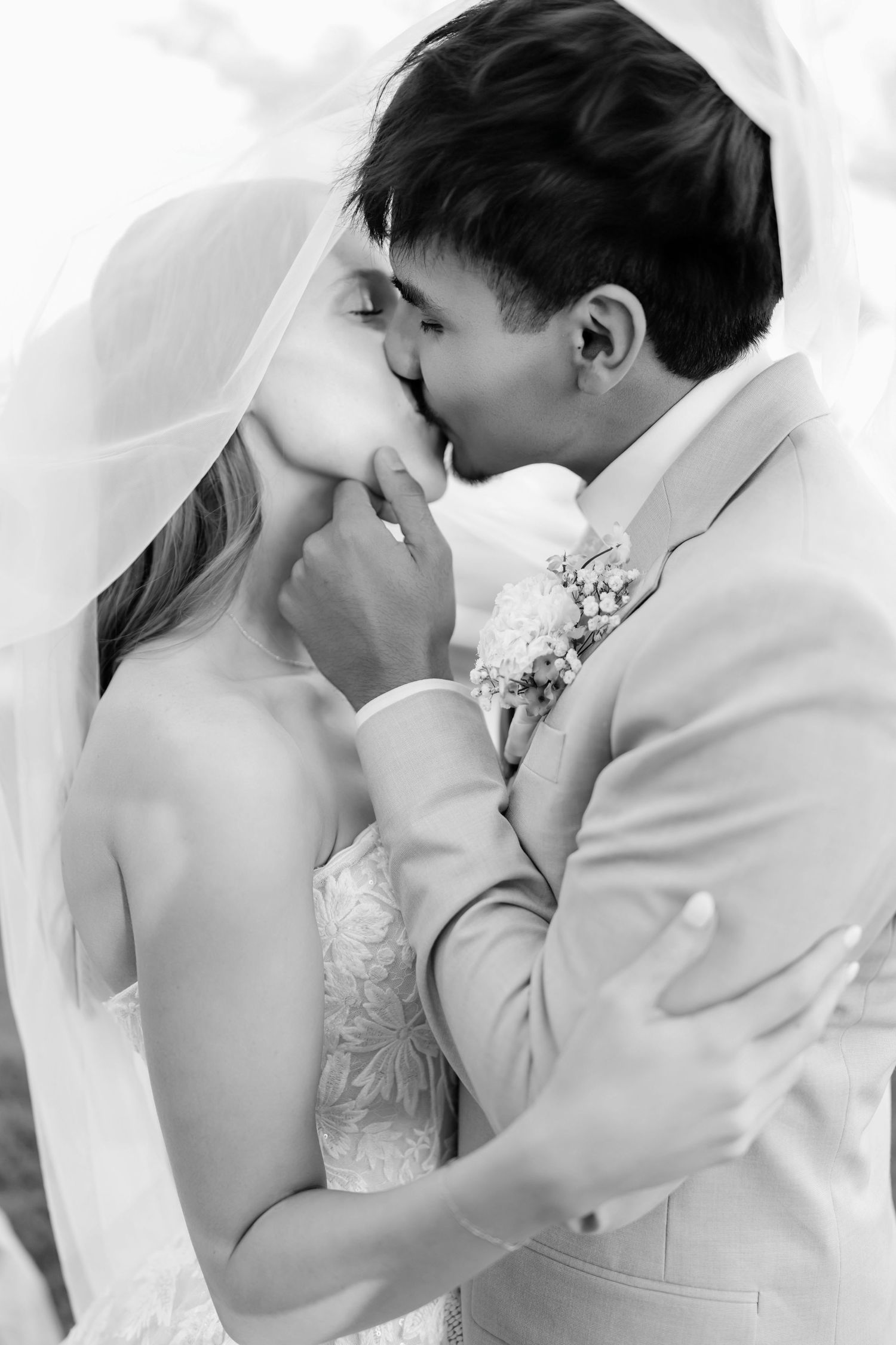 Black and white photo of a bride and groom sharing a romantic kiss under a flowing veil on their wedding day.