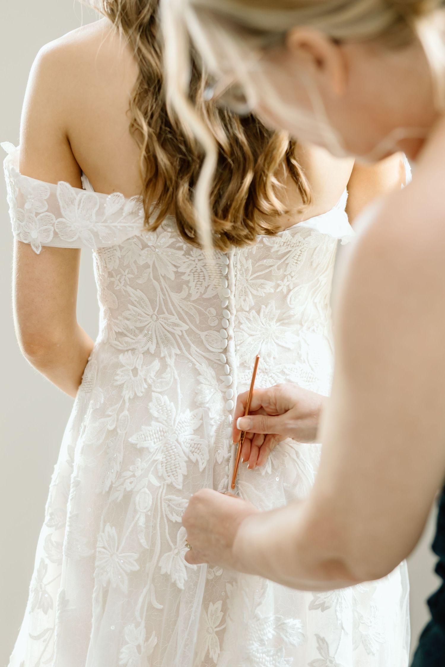 Bridesmaid fastening buttons on back of white lace off-shoulder wedding dress with floral embroidery.