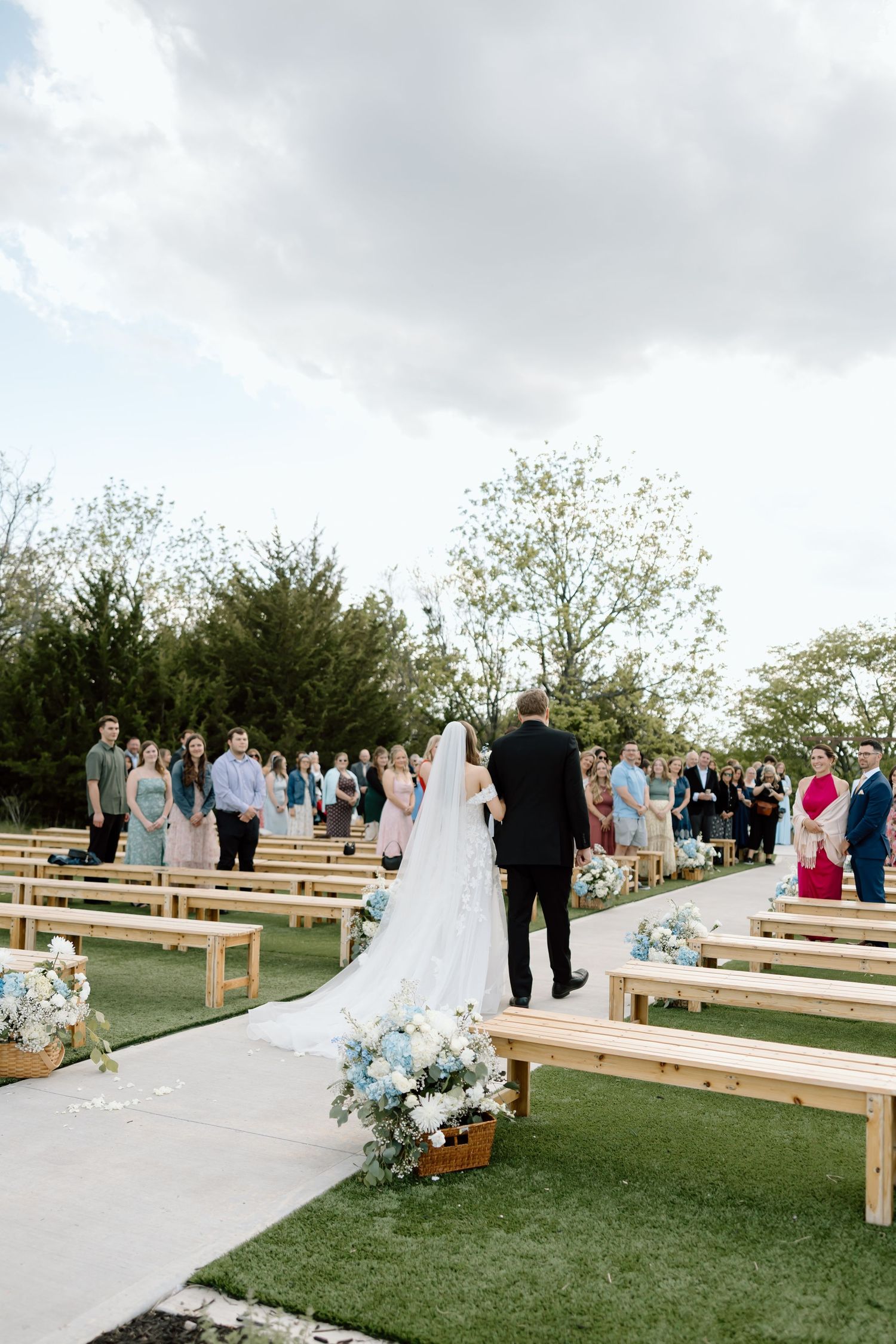 Bride walks down outdoor wedding aisle with wooden benches, blue floral arrangements, and guests standing under cloudy sky.