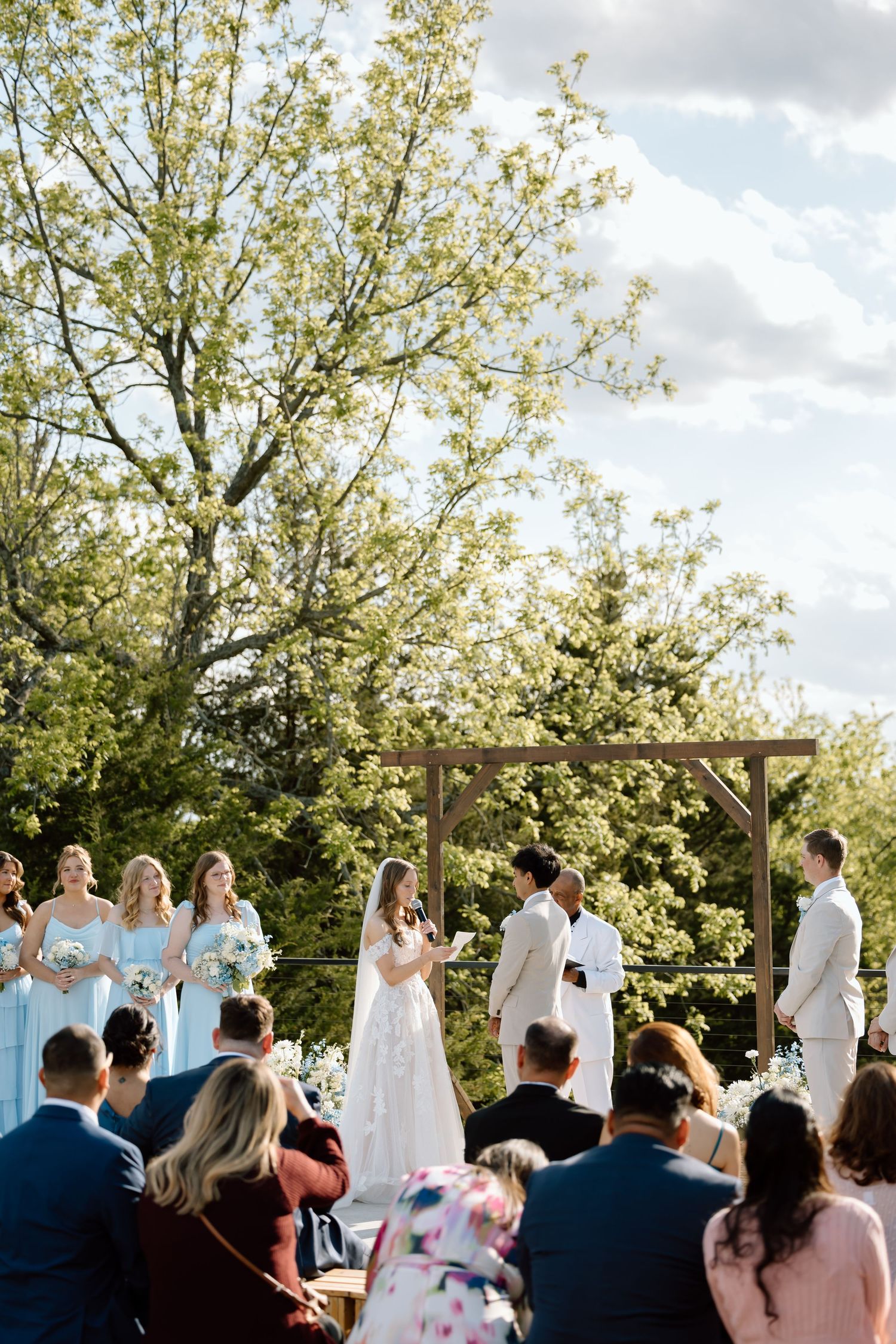 Outdoor wedding ceremony with bride and groom at wooden arch, bridesmaids in blue, guests seated on sunny day.