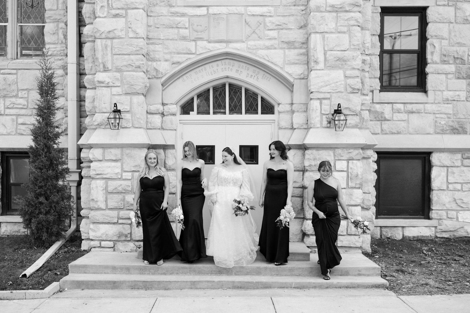 Bride in white gown poses with four bridesmaids in black dresses on stone church steps in elegant black and white photo.
