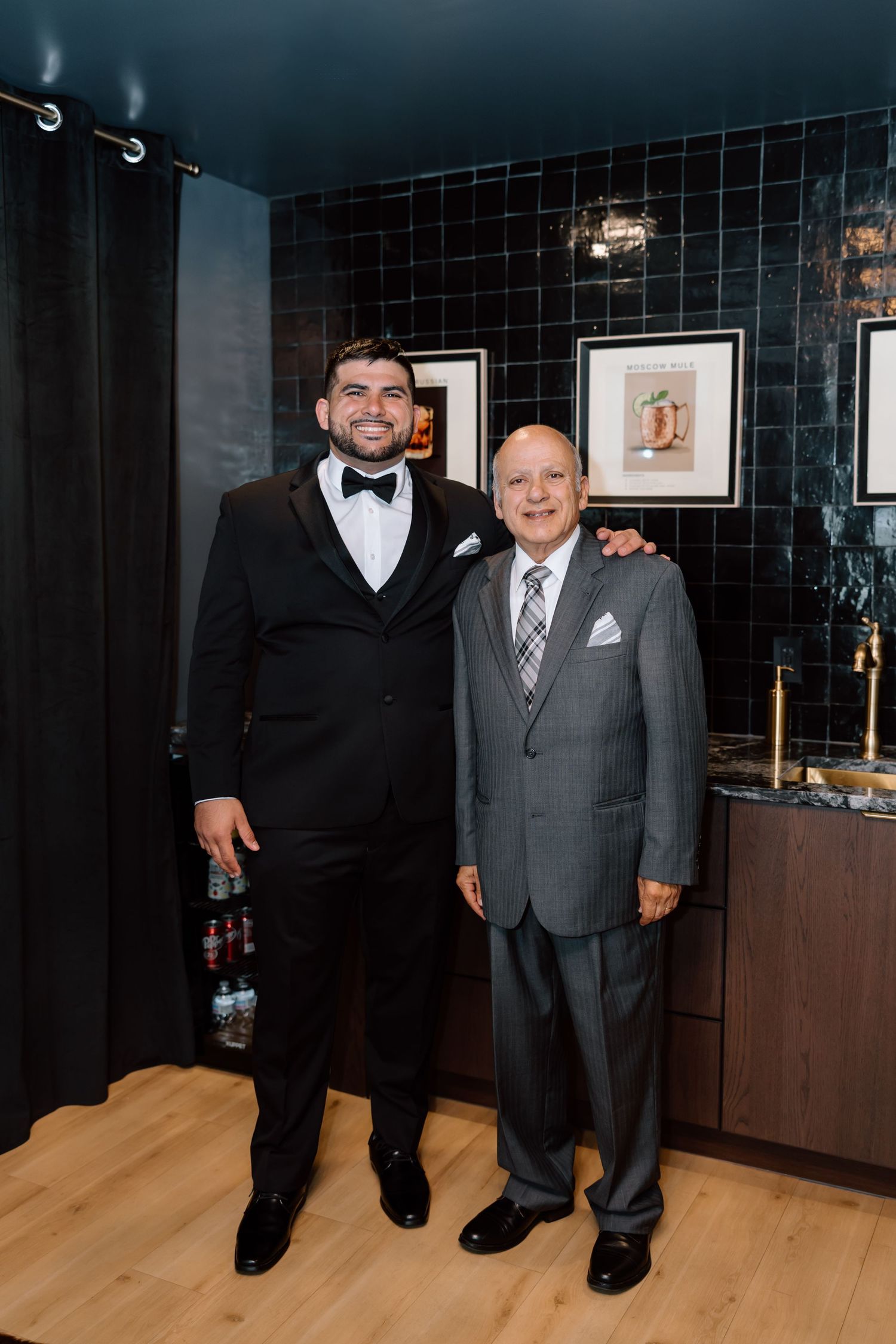 Groom in black tuxedo and bow tie posing with older man in gray suit in an elegant black-tiled dressing room.