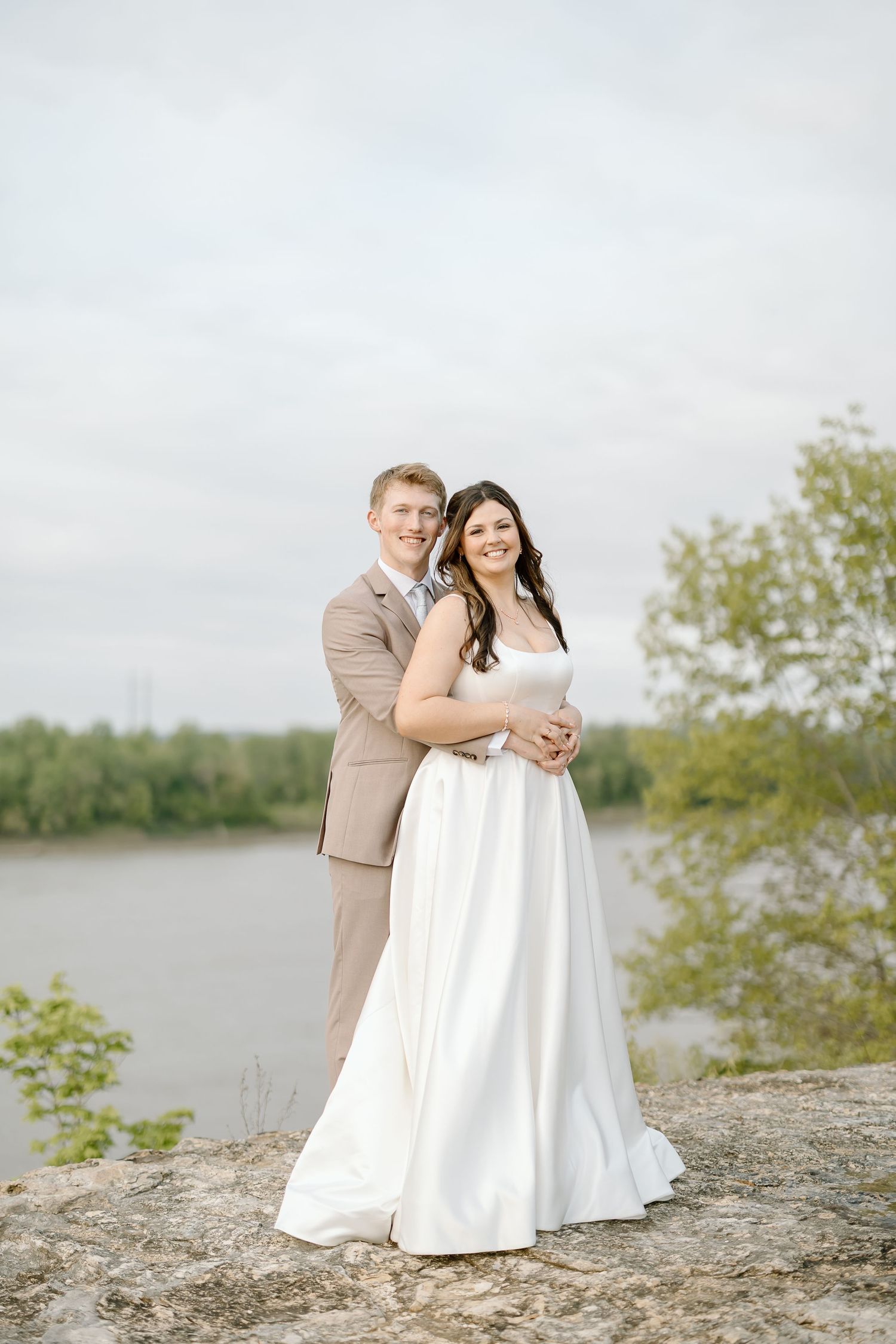 A romantic couple poses together on a rocky shoreline with a lake and trees in the background.