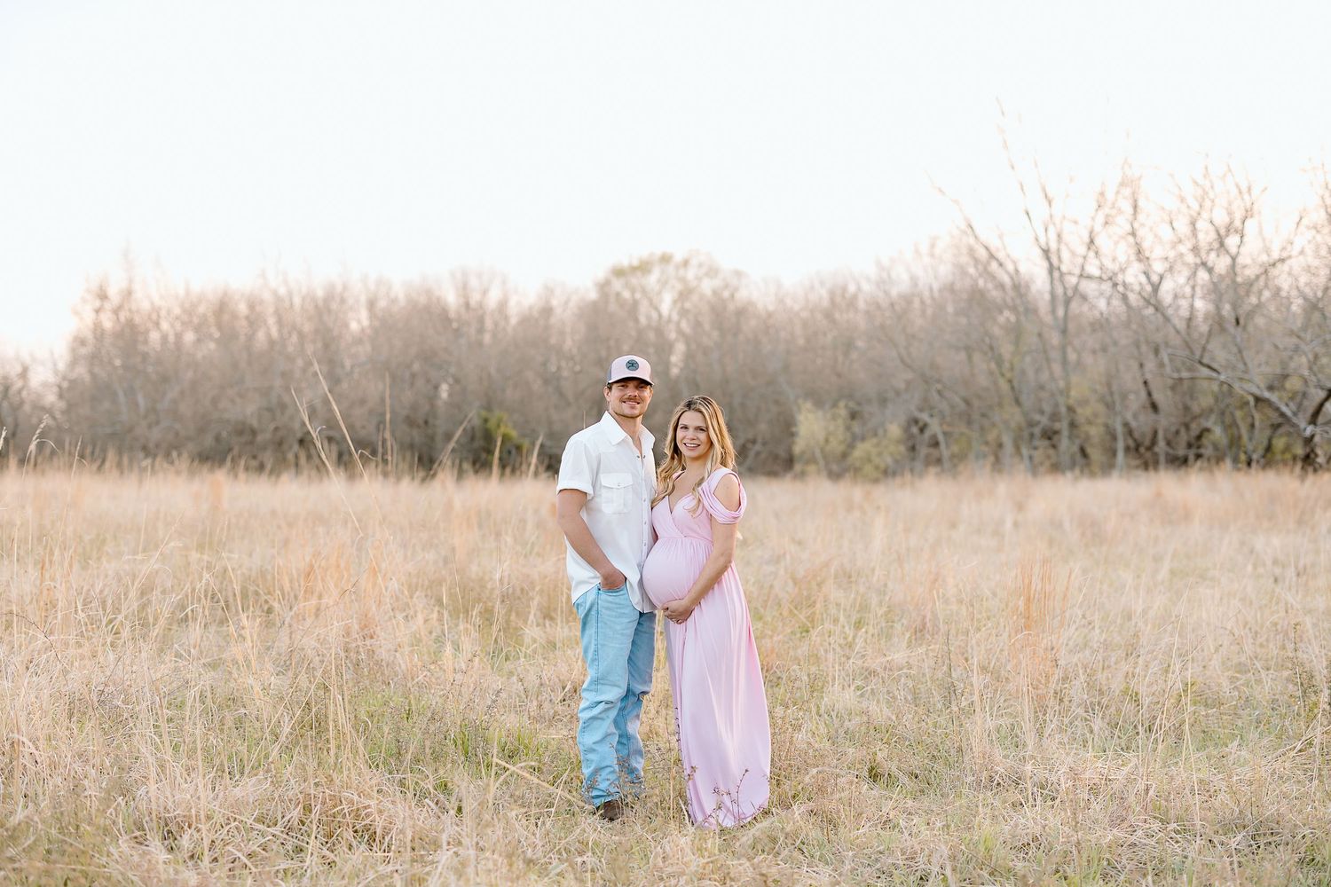 A couple in pastel outfits walks through a golden field during sunset with bare trees in the background.