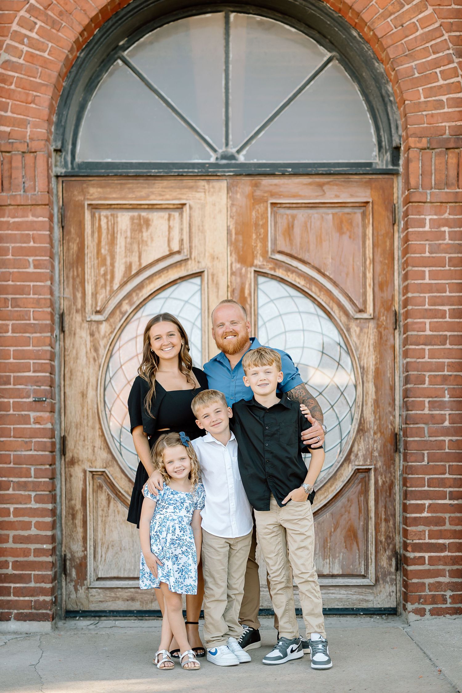 Three children stand together in front of rustic wooden church doors with brick archways.