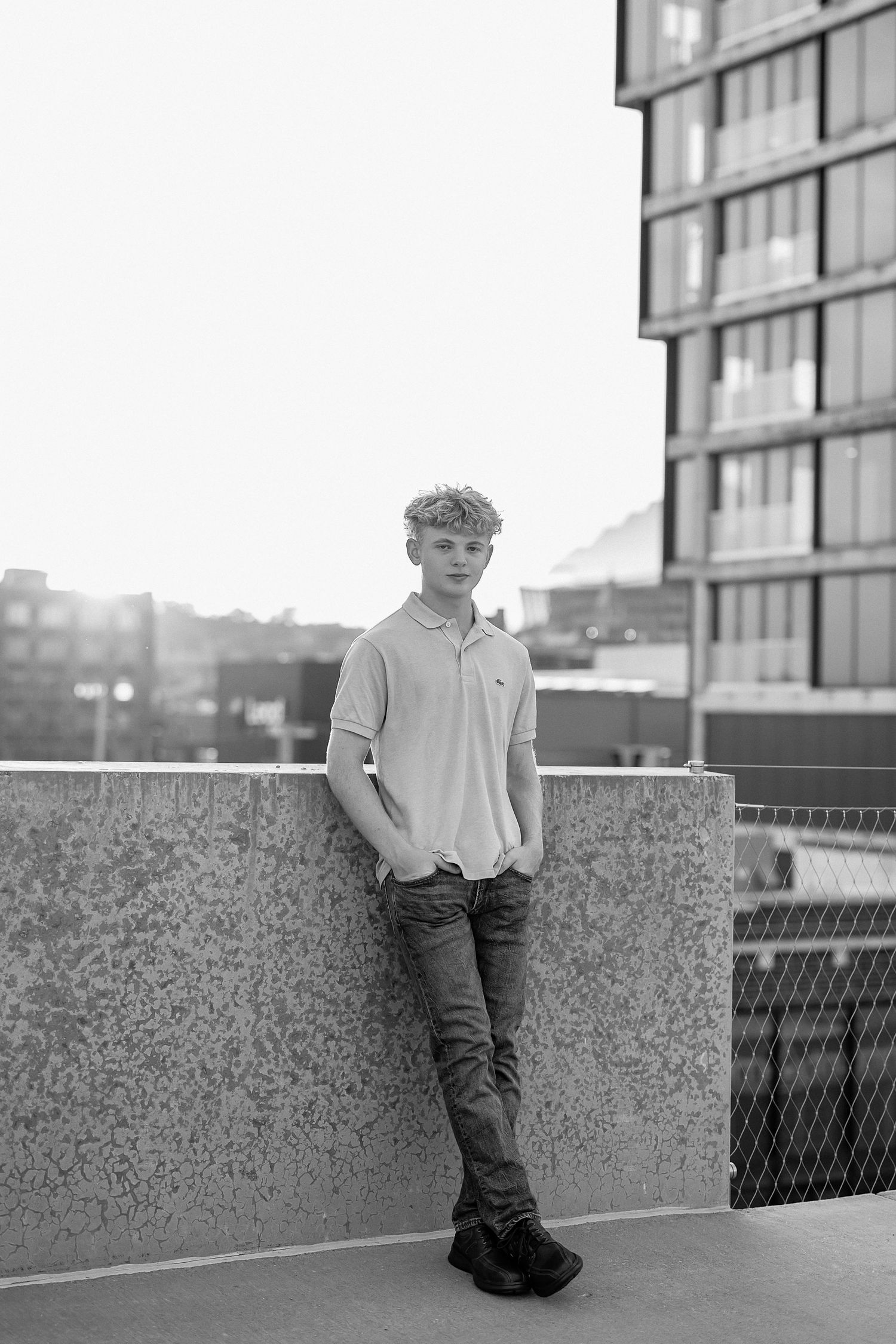 Black and white portrait of someone in casual attire leaning against a concrete wall on an urban rooftop.