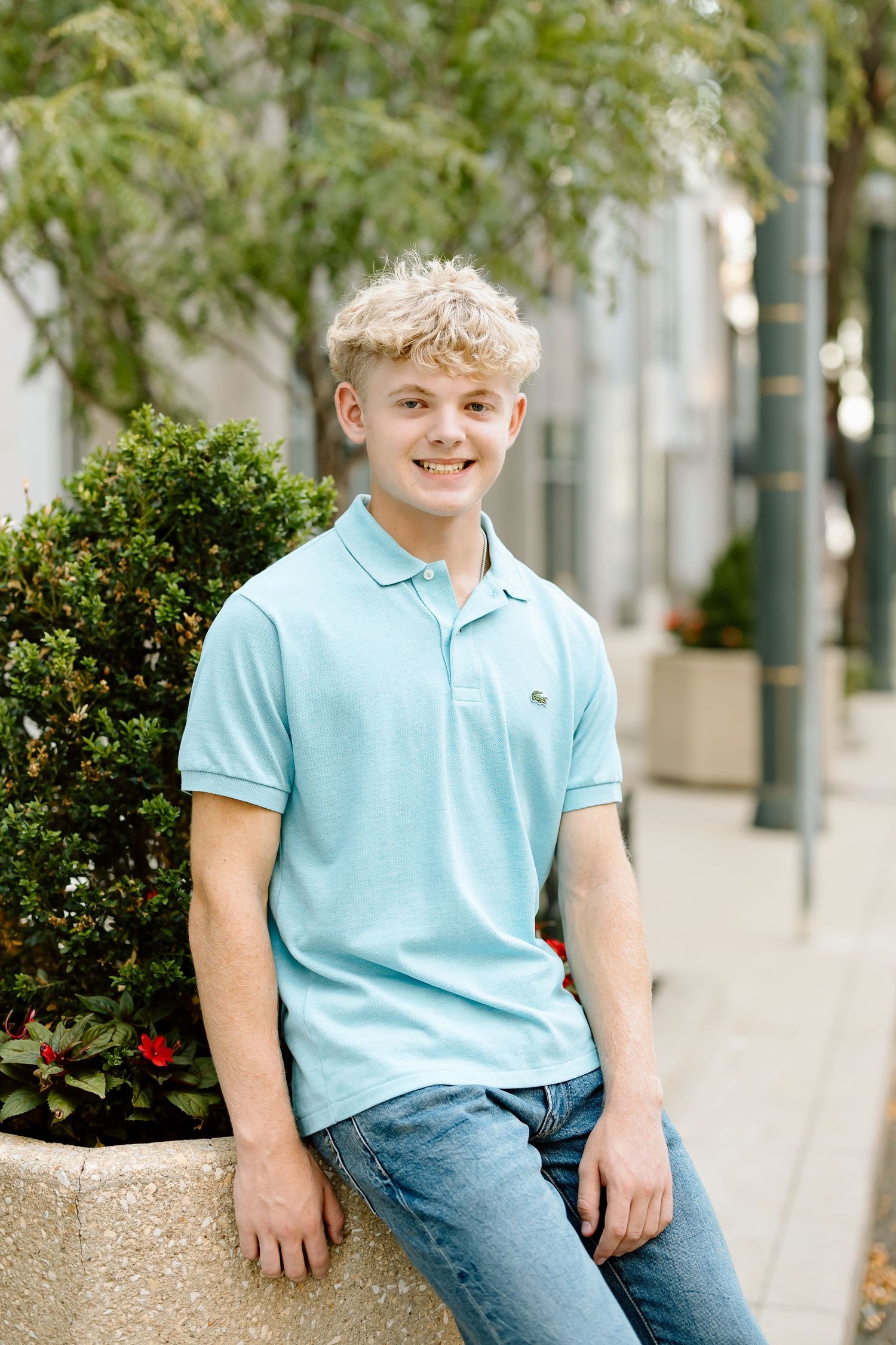 A young person in a light blue polo shirt and jeans sits casually on a stone ledge near landscaped bushes.