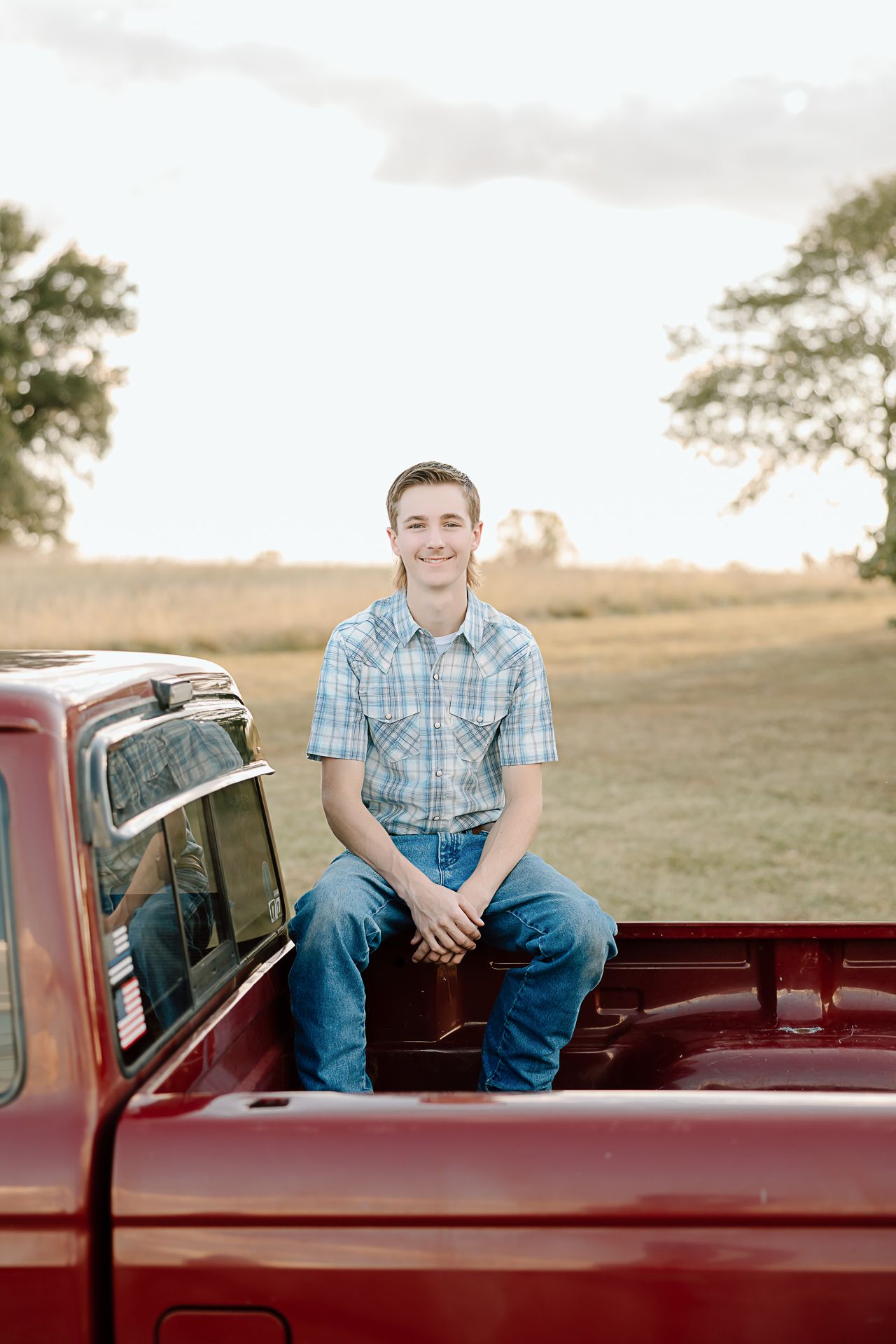 A casual portrait of someone sitting on the bed of a vintage red pickup truck in a rural field at sunset.