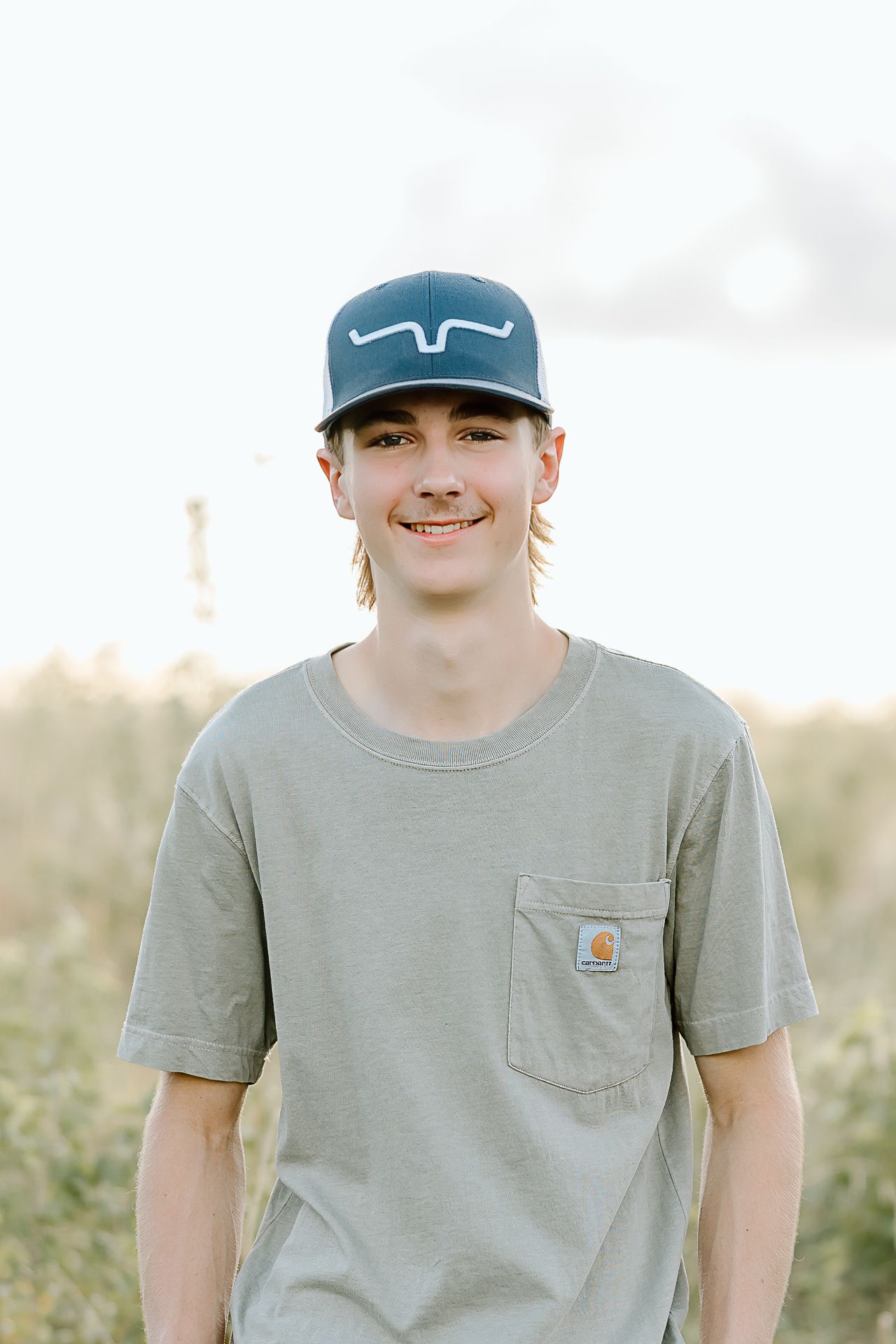 Young person wearing a gray Carhartt t-shirt and blue baseball cap stands outdoors in a field at sunset.