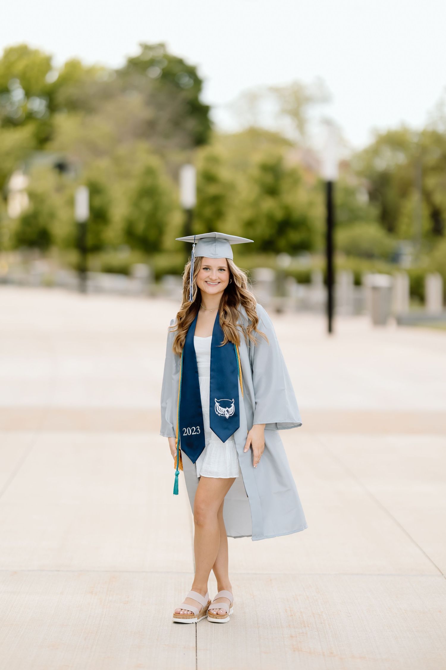 A graduate in a blue jean jacket and white dress walks along a sandy beach path during a sunny day.
