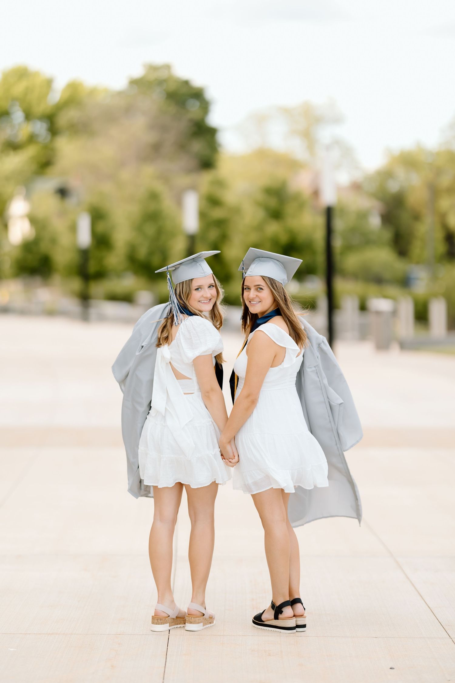 Two graduates in white dresses and grey graduation gowns pose together holding hands on a sunny day outdoors.