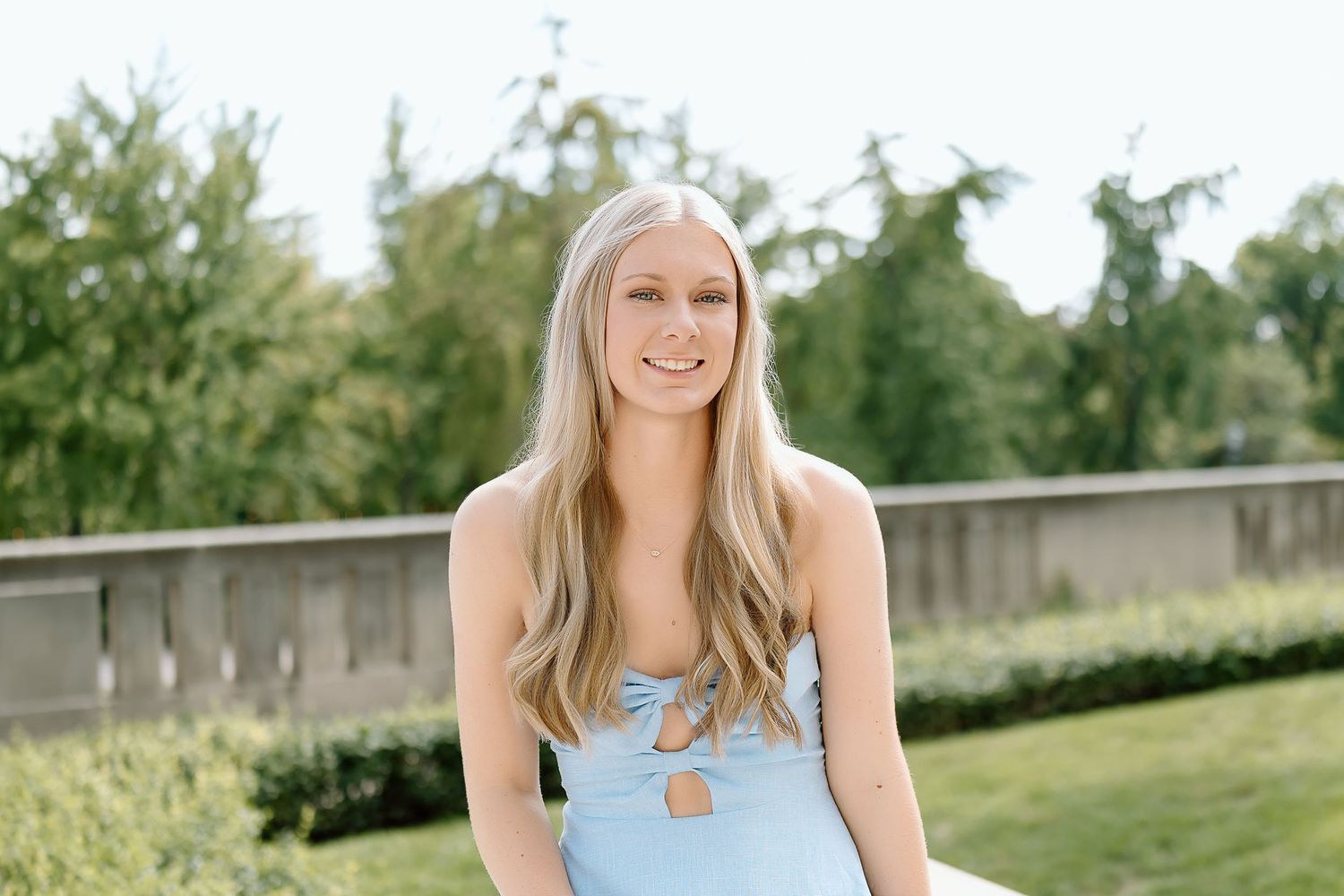 A person in a light blue sleeveless dress poses outdoors in a garden setting during a sunny day.