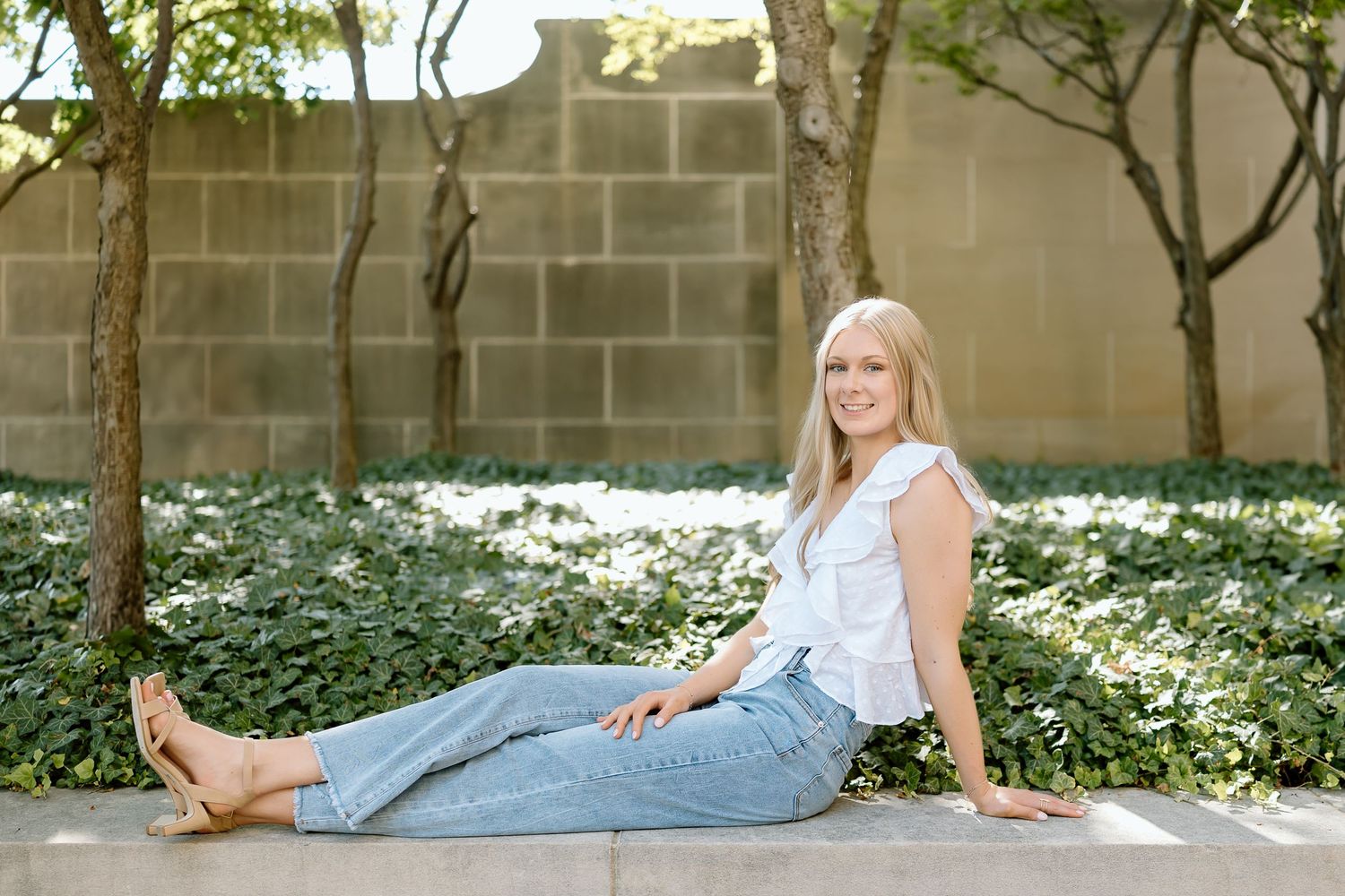 Person in light blue jeans and white sleeveless top sits casually on concrete ledge with flowering ground cover in background.