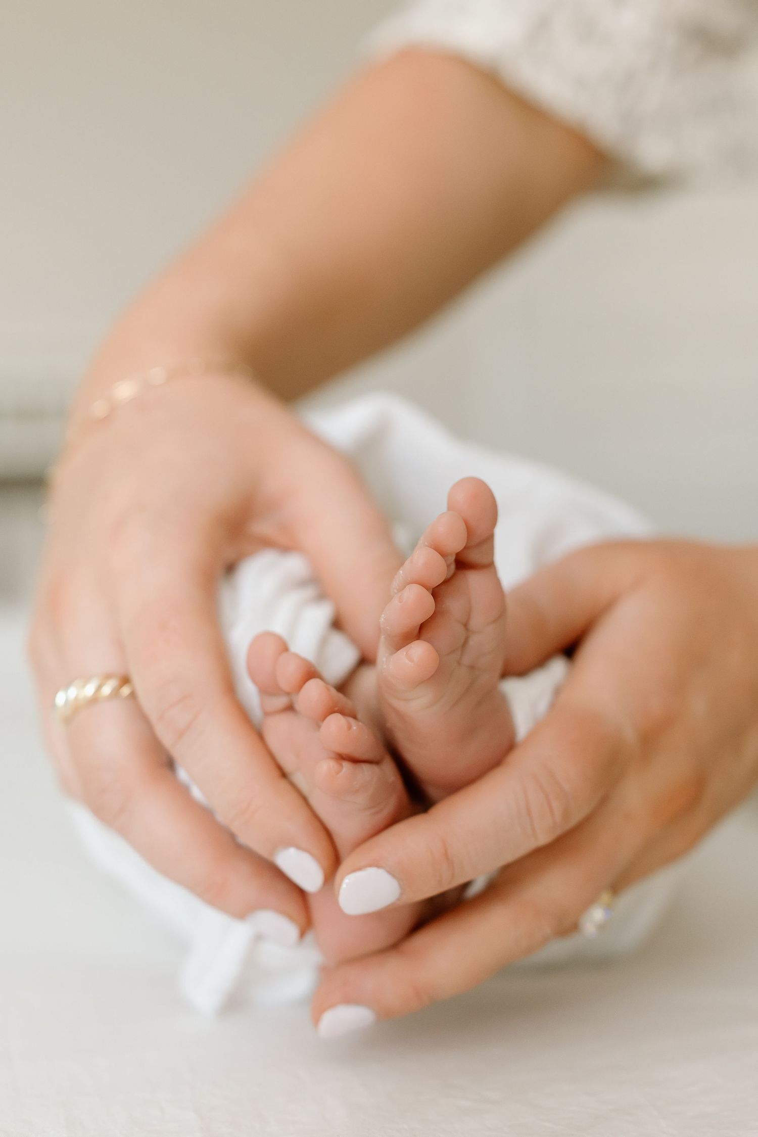 Adult hands with white manicured nails gently holding tiny baby feet against white fabric background.