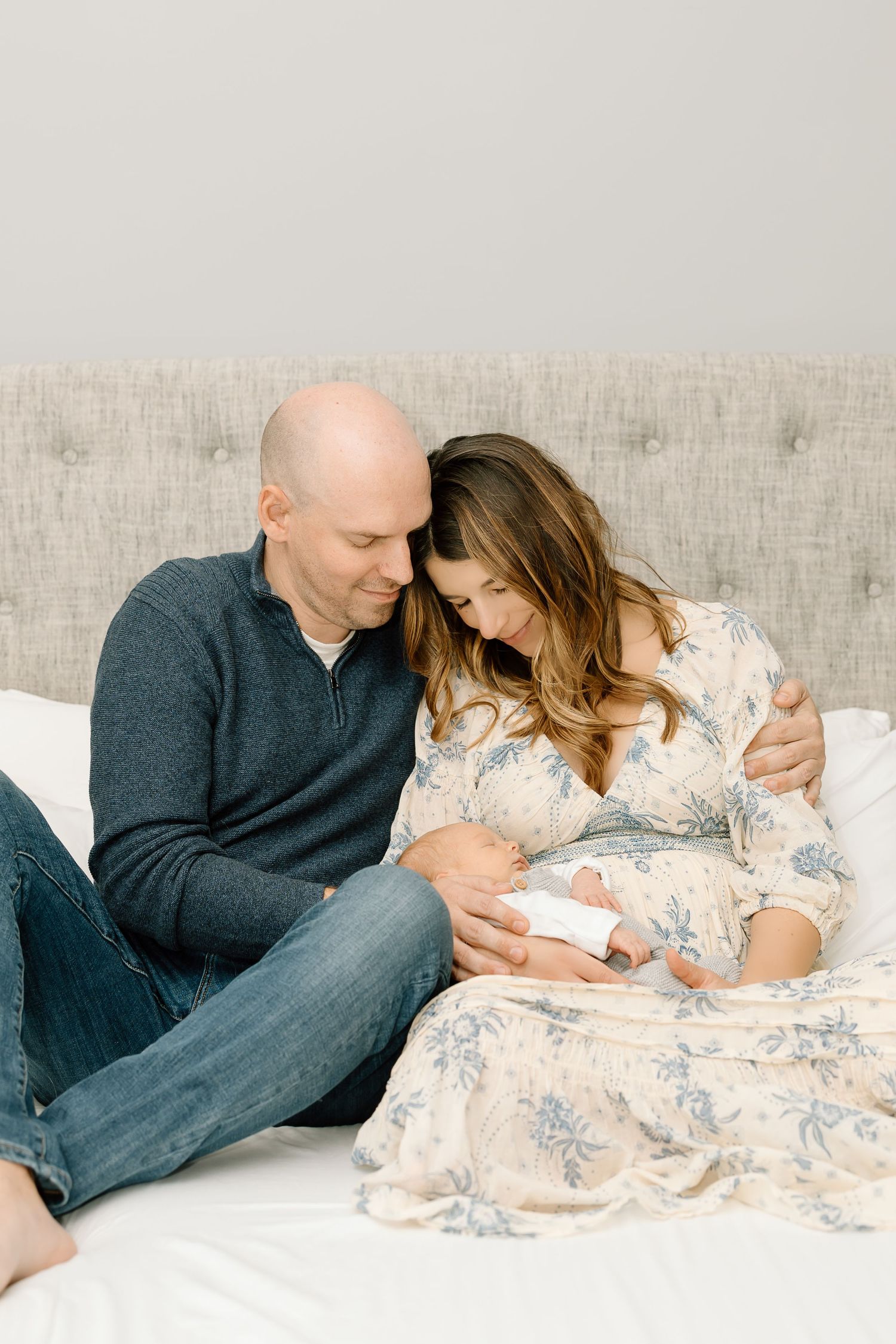A couple sits together on a white bed, sharing an intimate moment in casual attire against a gray backdrop.