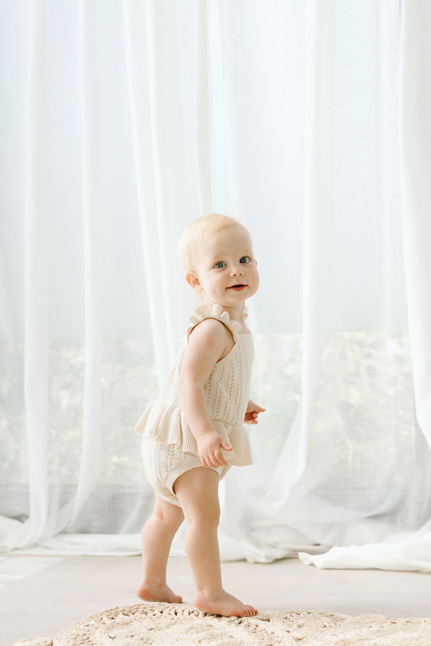 Baby in white onesie standing on textured rug near sheer curtains in bright natural light.