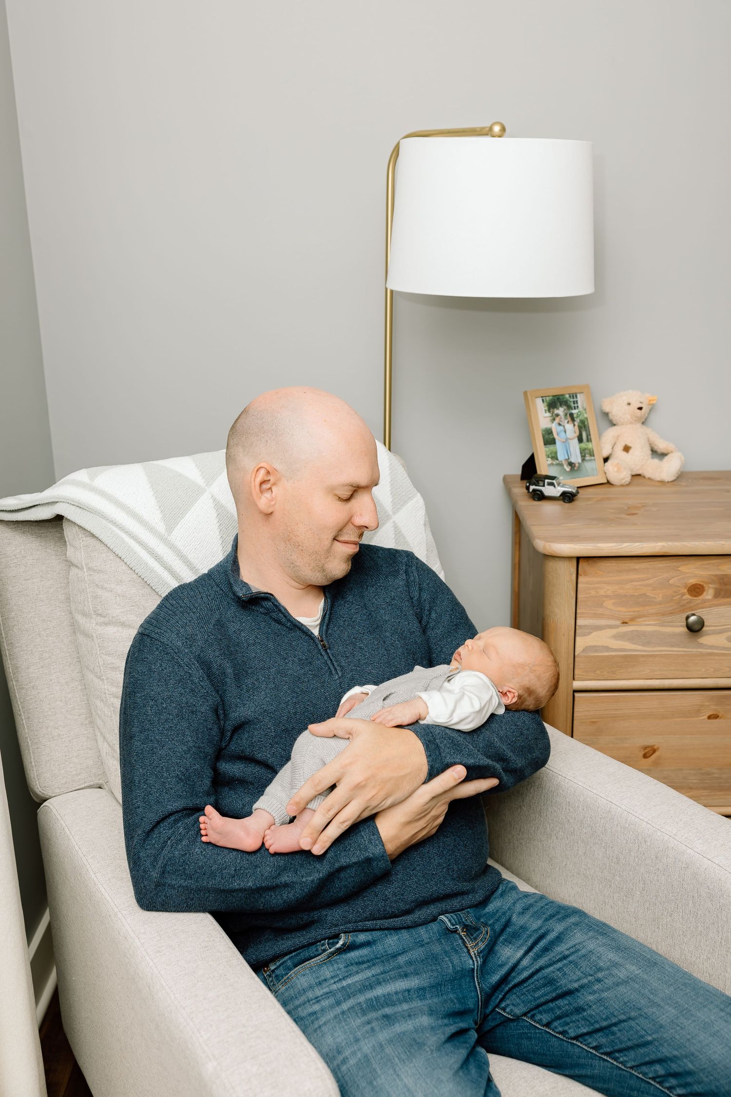 A person in a dark sweater sits in a cozy armchair holding and gazing lovingly at a sleeping newborn baby.