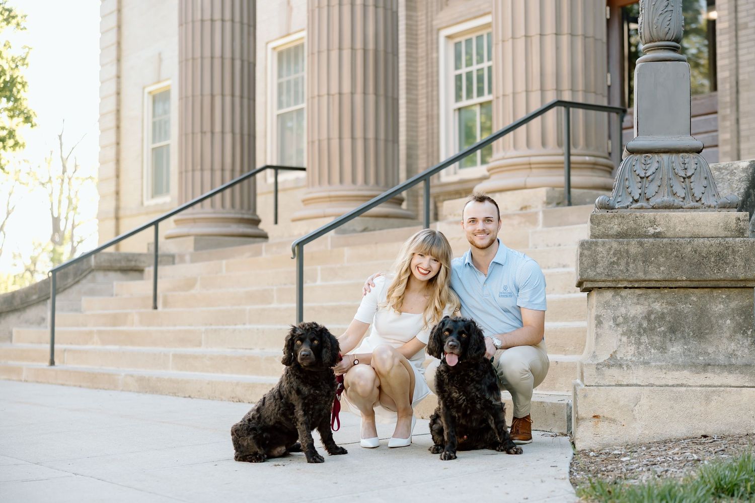 A couple sits on stone steps with two black dogs beside them at a historic building entrance.