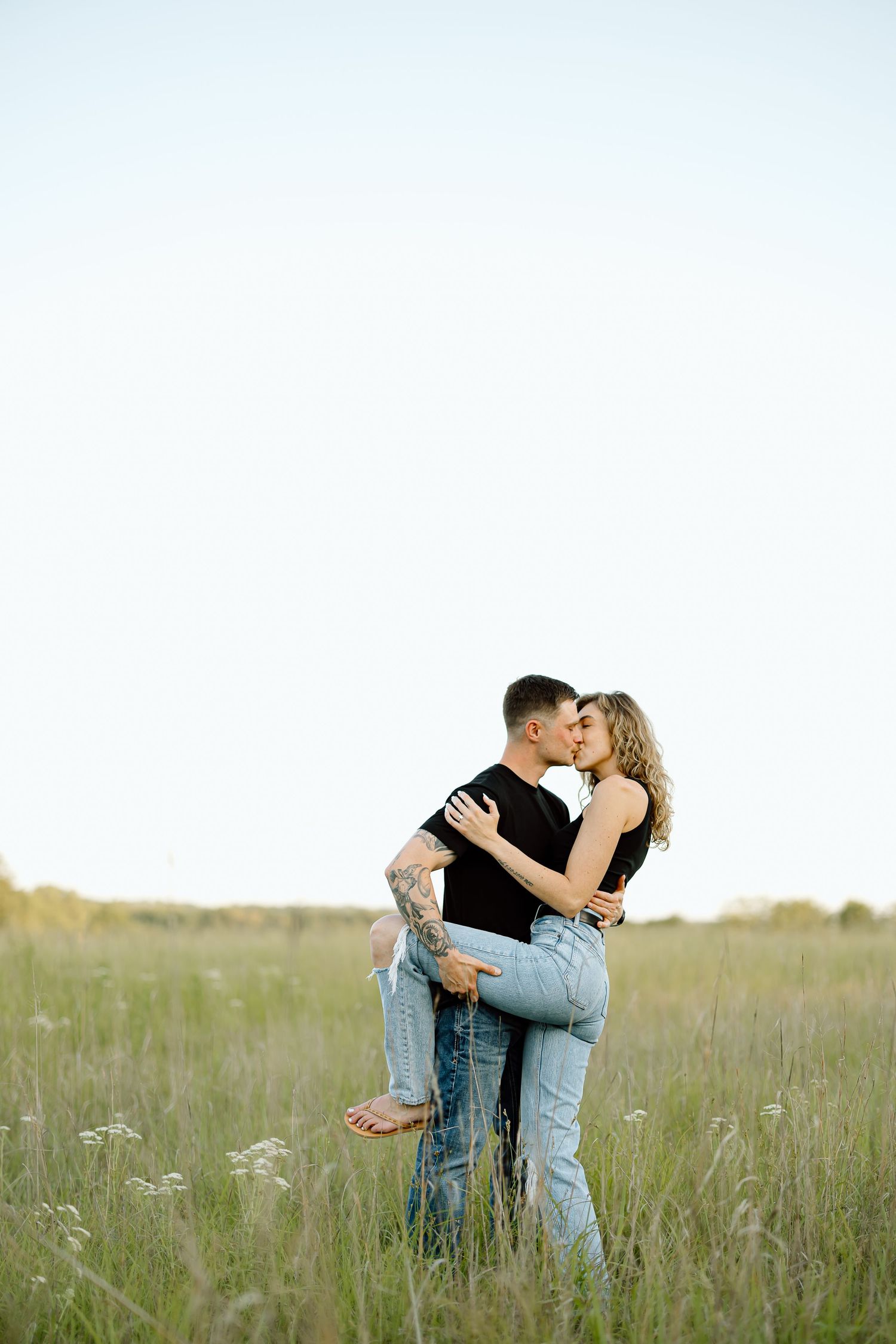 A romantic couple embraces and kisses in a serene meadow with tall grass and wildflowers at sunset.