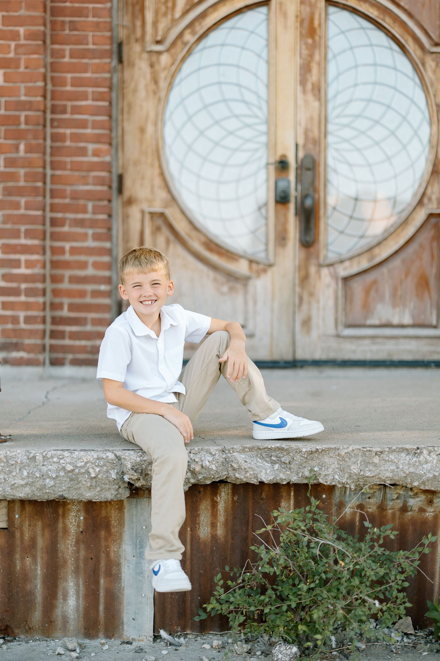Young boy in white shirt and khaki pants sits casually on concrete steps with ornate wooden doors in background.