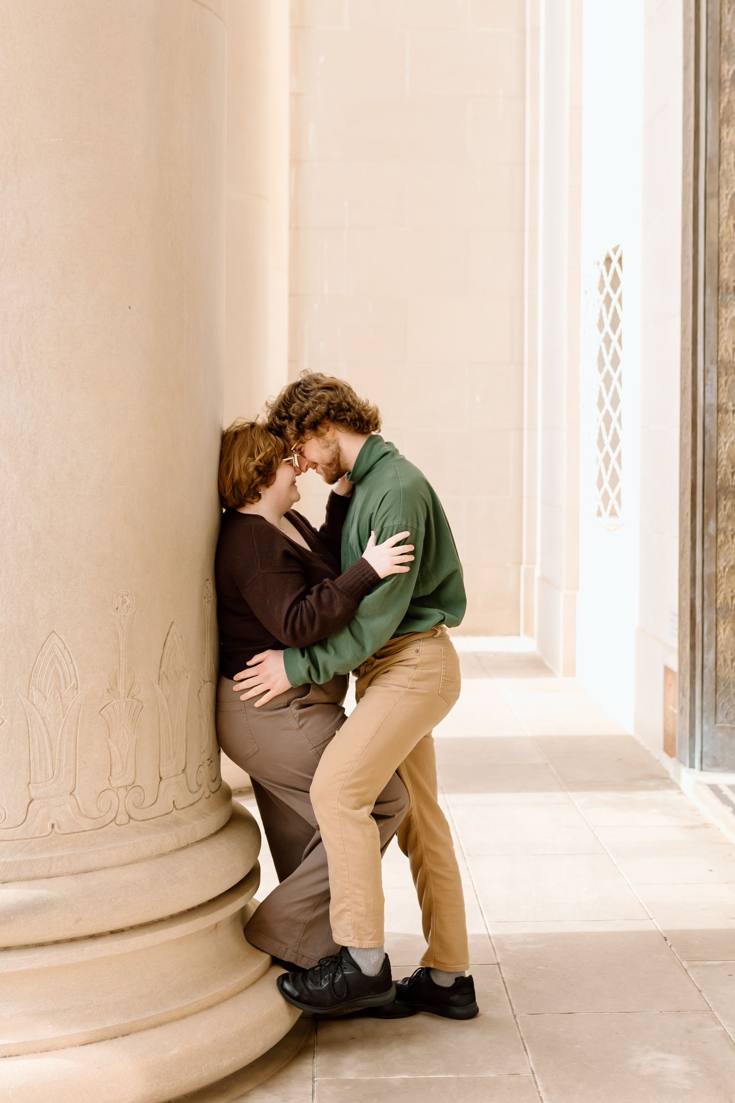 Two people embrace in a tender moment against a white architectural column in a minimalist setting.
