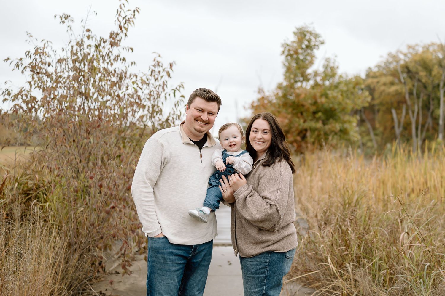Couple in cozy sweaters walking through autumn field with their small black dog on sunny afternoon.