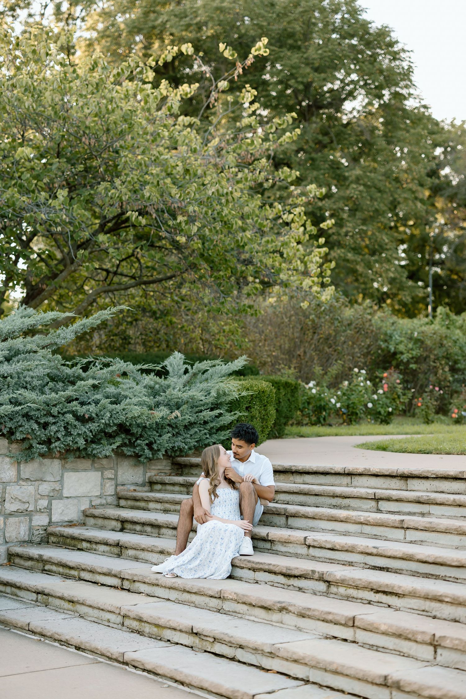A couple shares an intimate moment on stone amphitheater steps surrounded by lush greenery in a park setting.