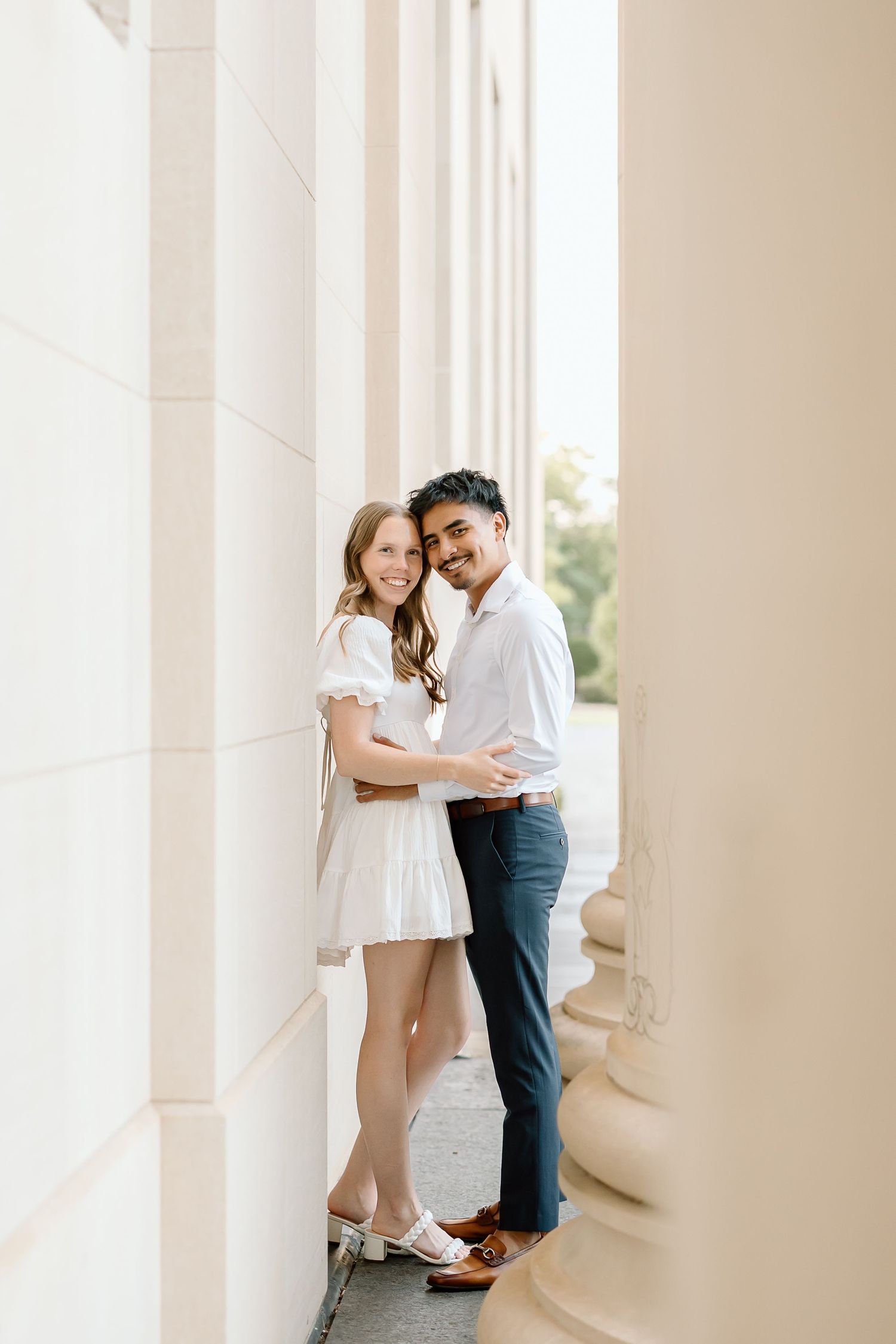 A romantic couple in white and blue attire embraces in an elegant columned hallway with soft natural lighting.