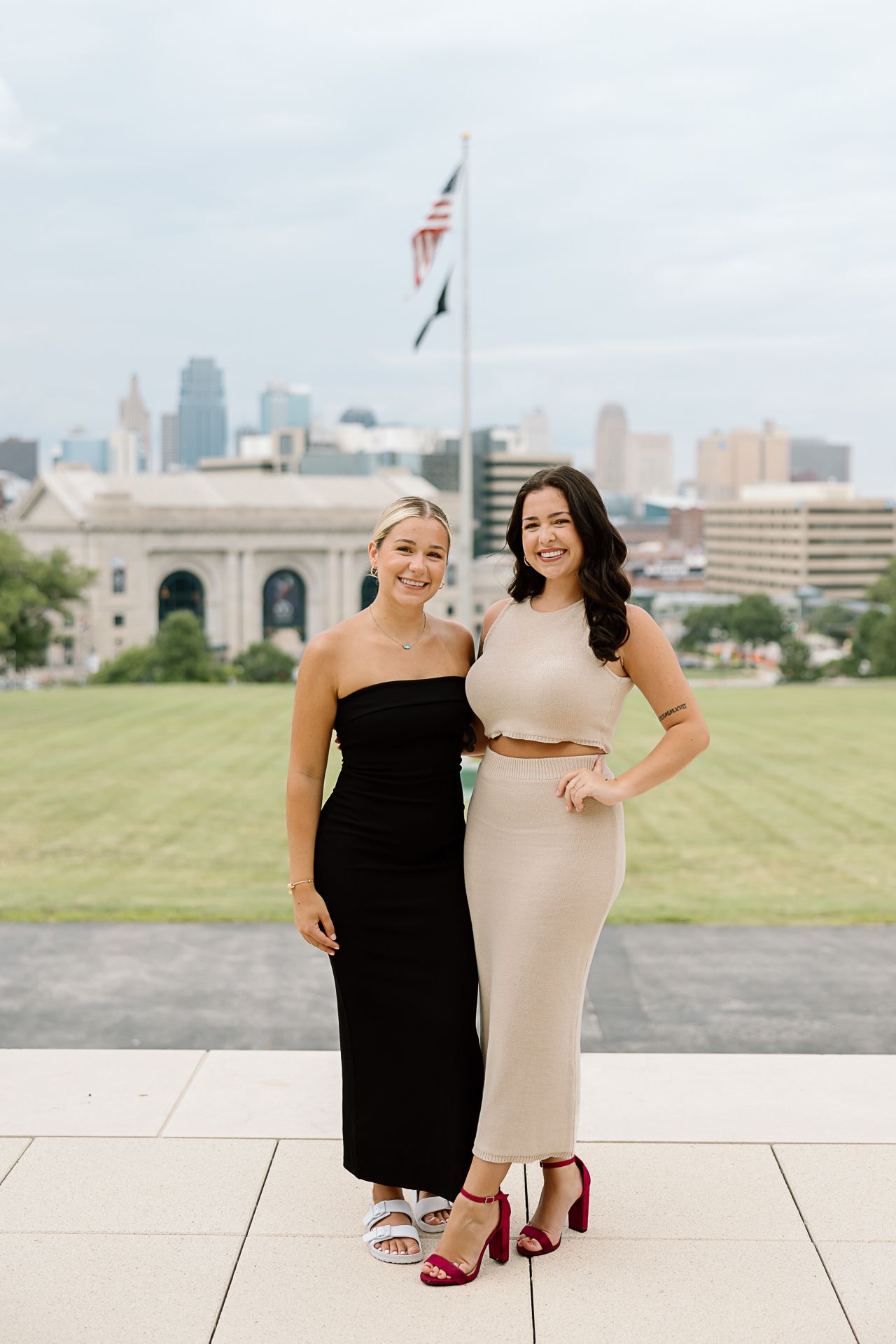 Two women in contrasting black and beige dresses pose together on a paved terrace with a city skyline backdrop.
