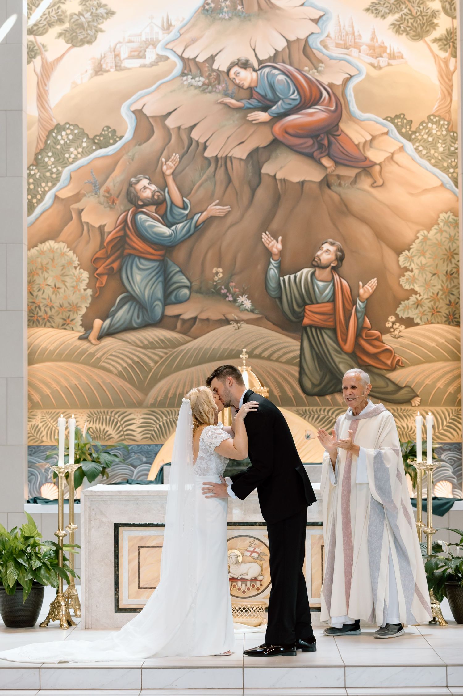 Bride and groom share first kiss at church altar with religious mural backdrop, officiant standing nearby.