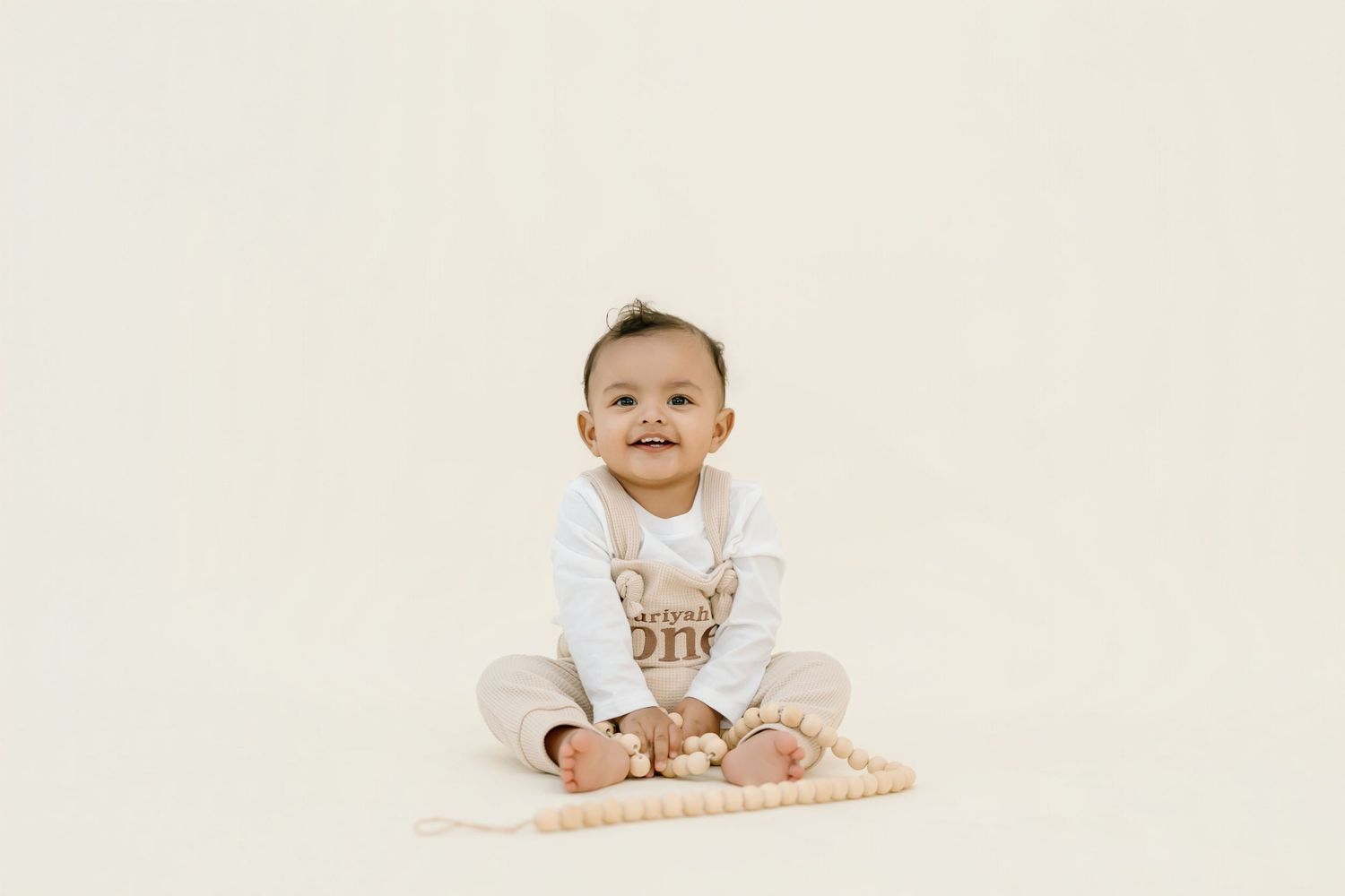 Baby wearing white outfit sits on cream background with a happy expression.