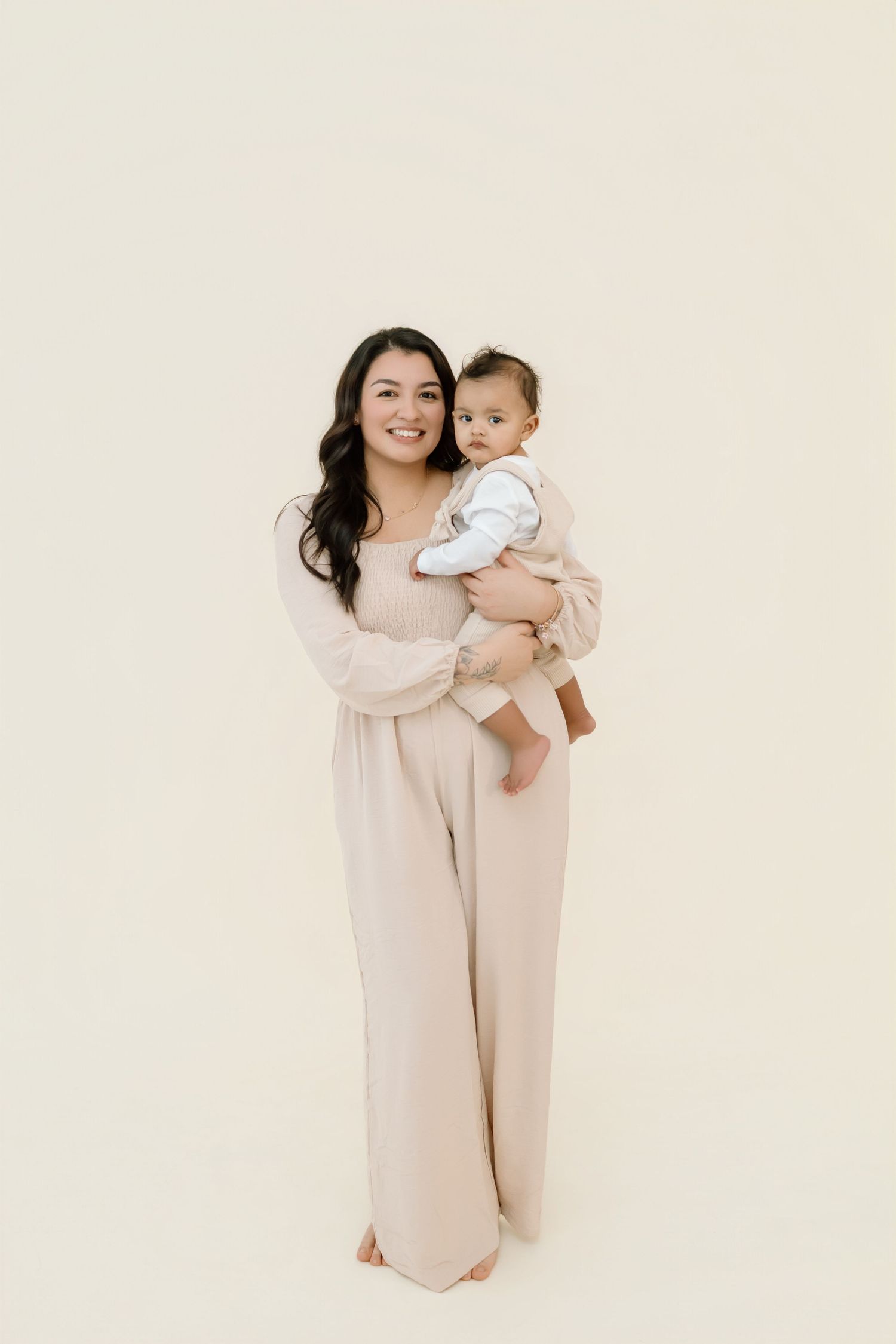 A parent and baby wearing matching cream-colored outfits pose together for a series of studio portraits.