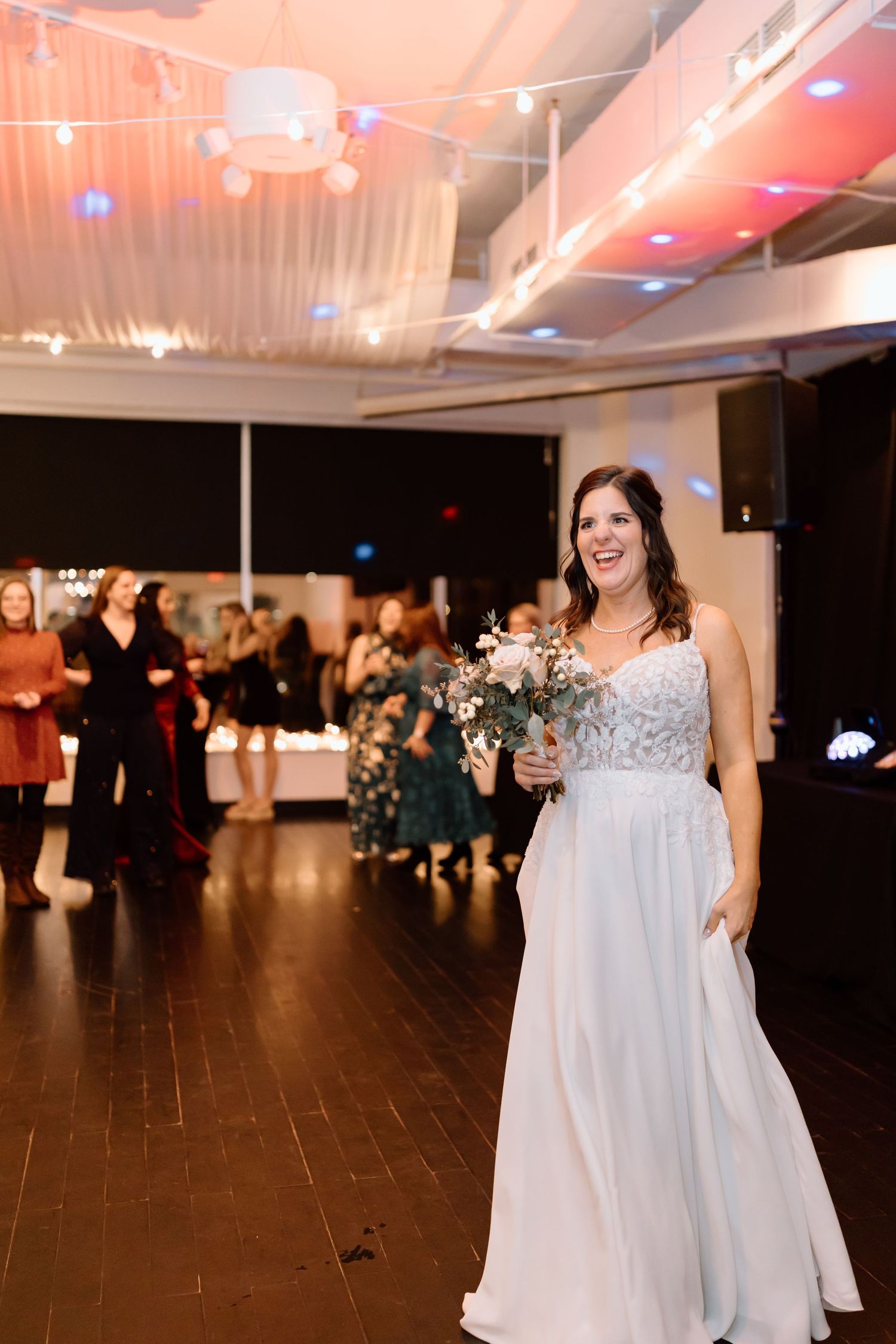 A group of people dancing and celebrating at a wedding reception with string lights and white decor.