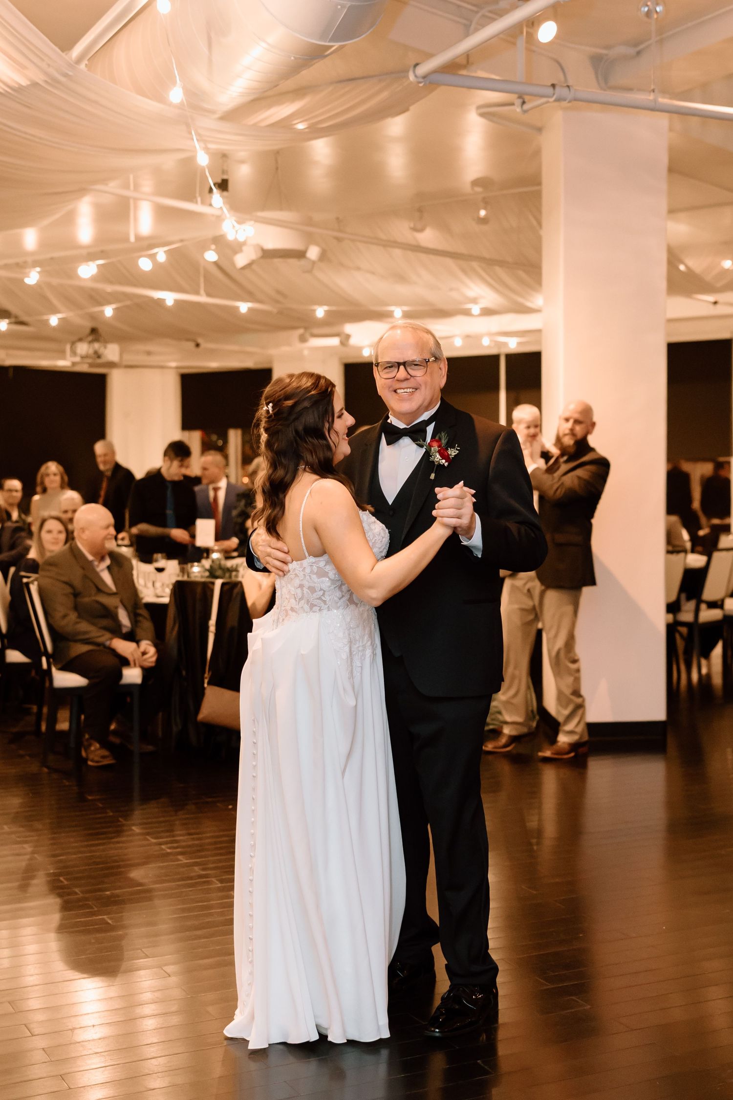 Couples dance together on a polished wooden dance floor at an elegant indoor reception with white columns and string lights.