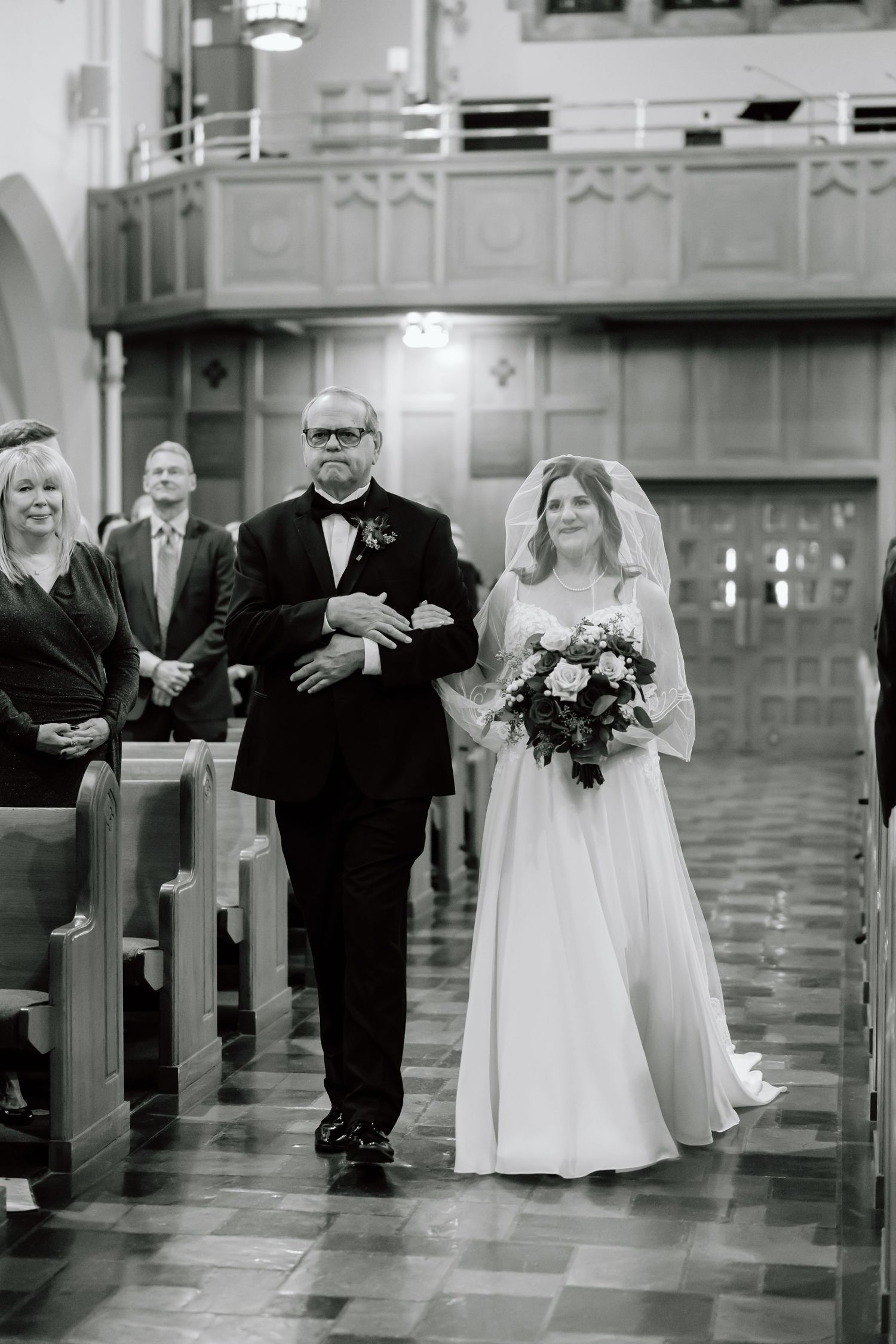 A black and white wedding photograph showing a bride in white dress walking down the church aisle during ceremony.