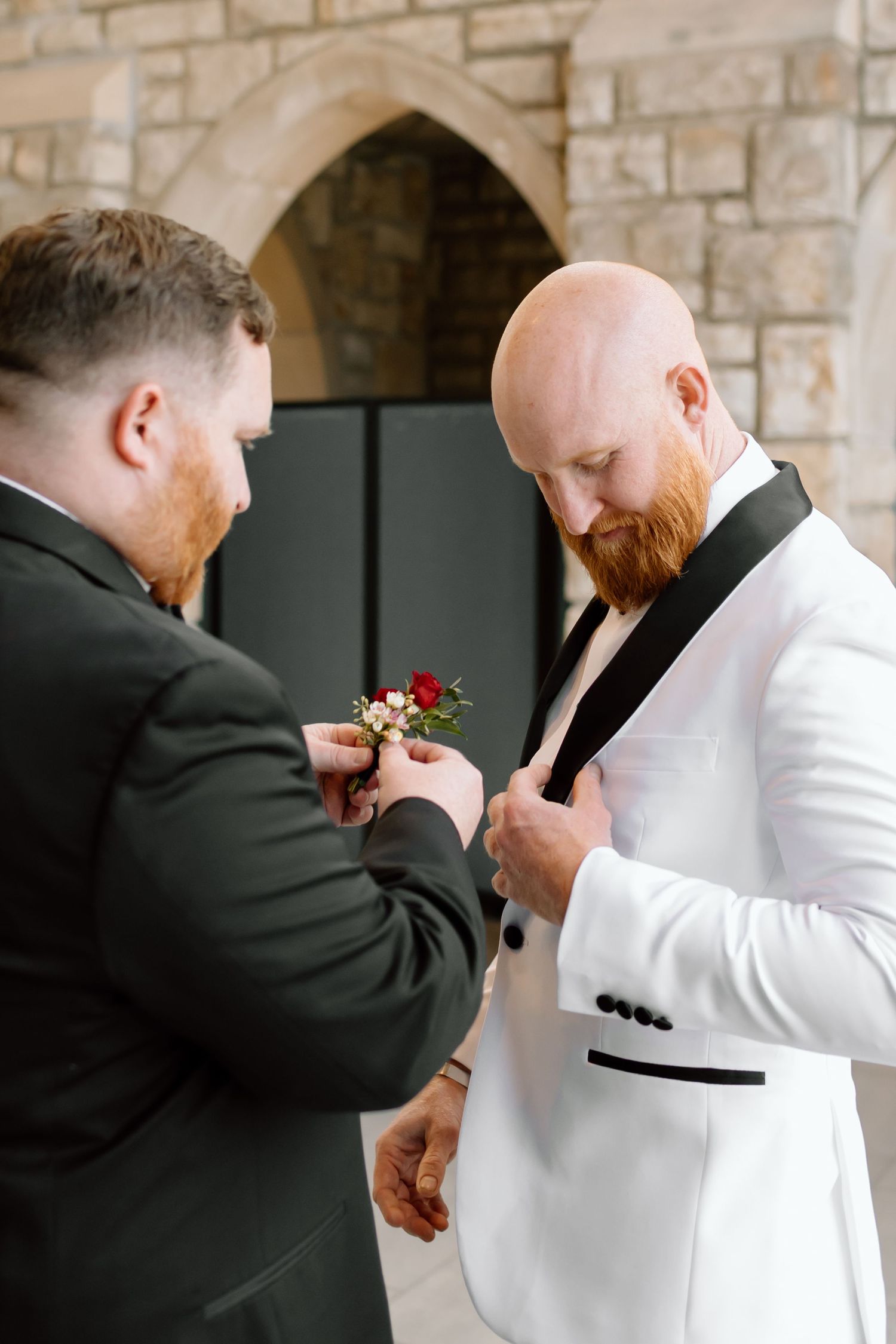 Two men in formal attire help each other with boutonniere flowers during a wedding preparation.