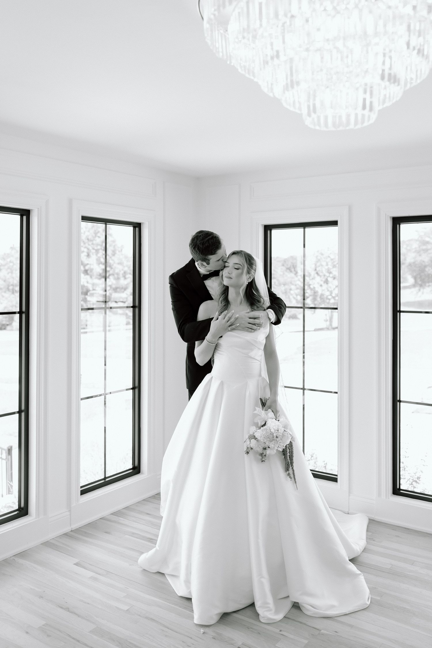 Black and white photo of a groom embracing his bride in a white gown, holding a bouquet, in an elegant windowed room.