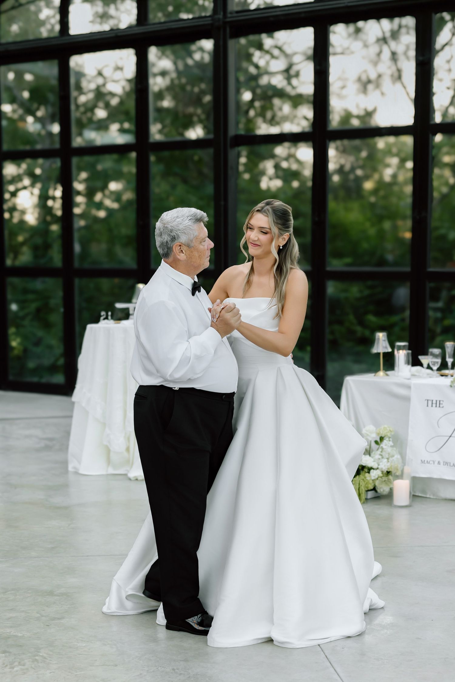 Bride in white ball gown shares father-daughter dance at elegant wedding reception with large windows.