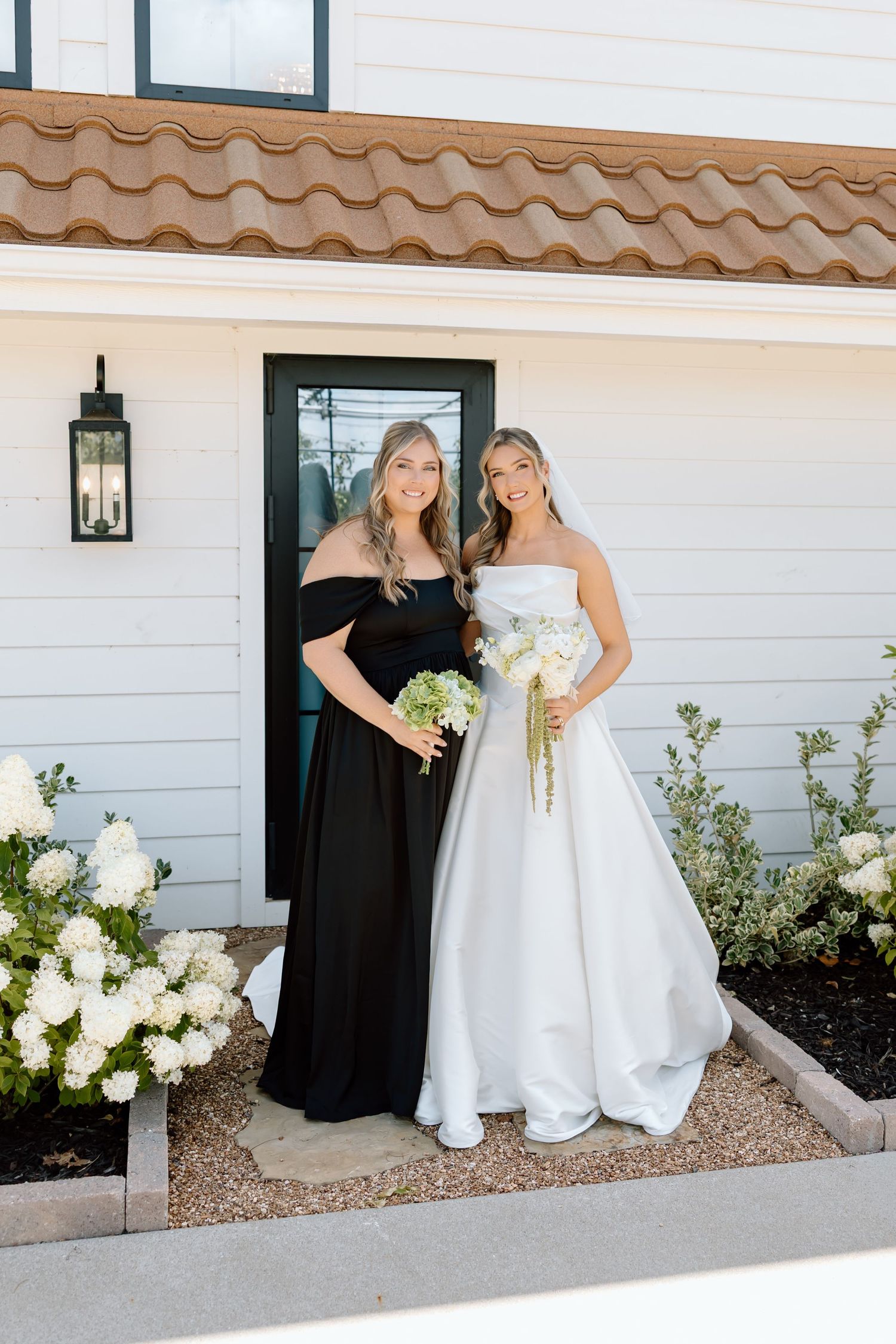Bride in white strapless ball gown and bridesmaid in black off-shoulder dress holding bouquets outside white home.